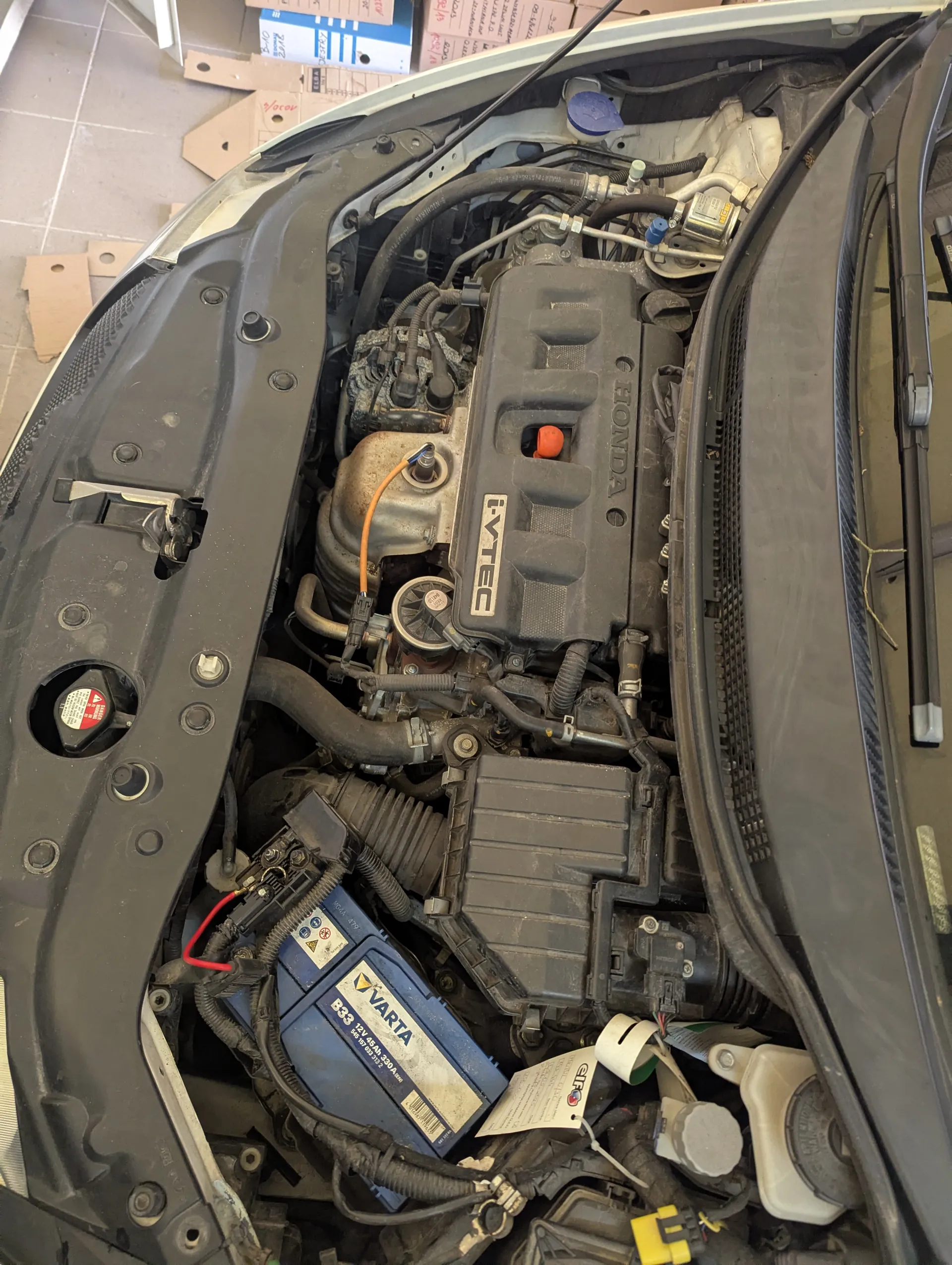 Widok otwartej tylnej części silnika samochodu marki Honda z widocznym napisem HONDA i i-VTEC na pokrywie silnika. W dolnej części obrazu znajduje się niebieski akumulator VARTA B33 z etykietą. Wokół silnika widoczne są przewody, węże oraz różne elementy mechaniczne. W tle na podłodze leżą pudełka i kartony. Całość sfotografowana z góry, z otwartą maską samochodu.