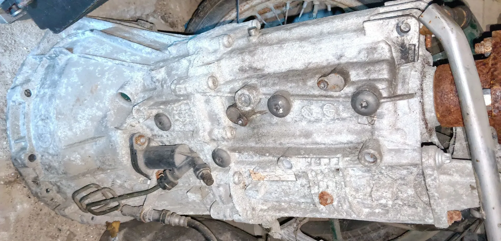Zbliżenie na metalową obudowę skrzyni biegów samochodu. Element jest srebrny, pokryty osadem i zabrudzeniami. Widoczne są liczne śruby, otwory montażowe oraz przewody hydrauliczne i elektryczne podłączone do obudowy. Po prawej stronie widoczny jest fragment zardzewiałego elementu metalowego oraz srebrna rurka.