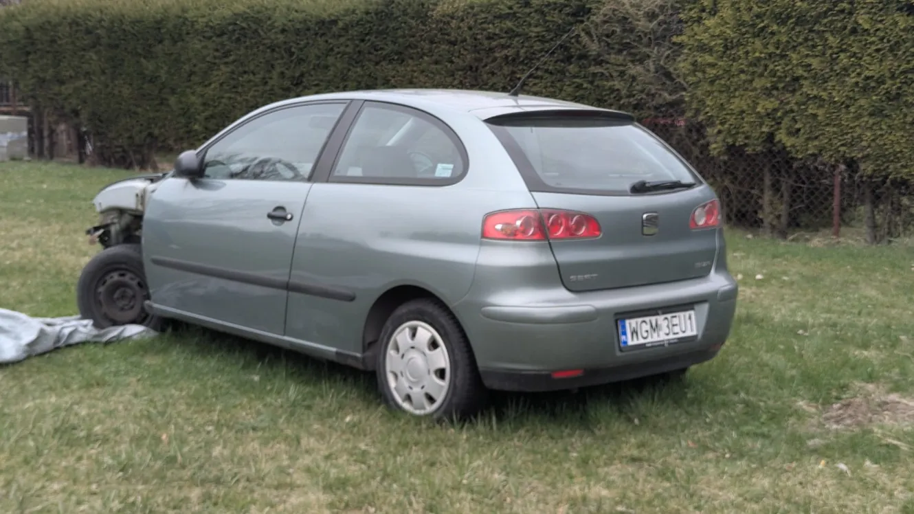 Szary samochód osobowy marki SEAT z widocznym uszkodzeniem przedniej lewej części, w tym brakującym kołem i otwartą maską. Na tylnej szybie widoczny znak rejestracyjny WGM 3EU1. Pojazd stoi na trawie, w tle zielony żywopłot i siatka ogrodzeniowa. Przy przodzie samochodu leży szara tkanina.