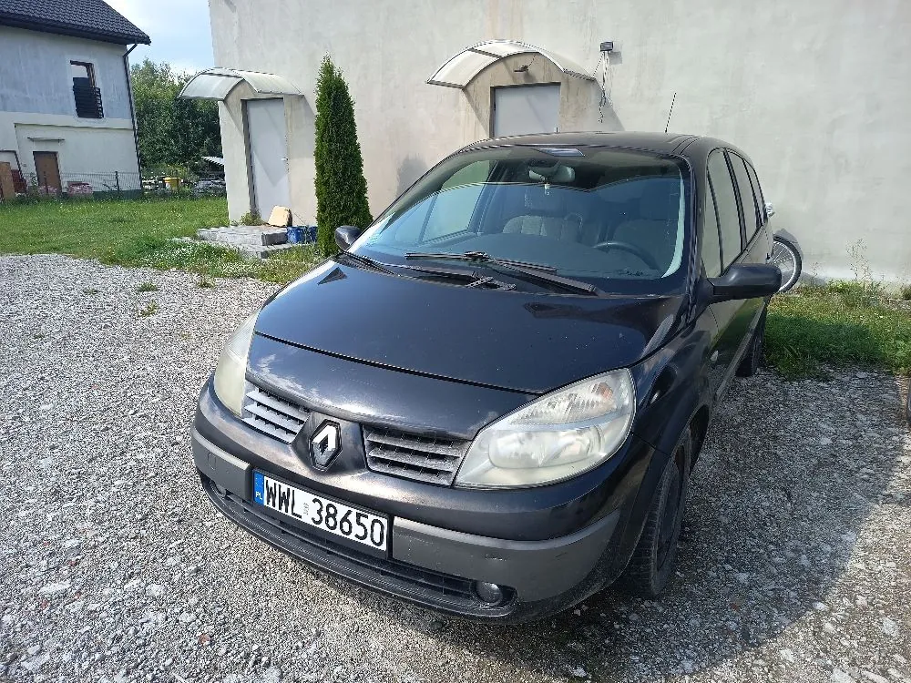 Czarny samochód marki Renault zaparkowany na żwirowym podjeździe przed jasnym budynkiem. Na przodzie pojazdu widoczna jest polska tablica rejestracyjna o numerze WWL 38650. W tle znajduje się budynek z dwiema białymi markizami nad wejściami oraz wysoki, wąski zielony krzew.
