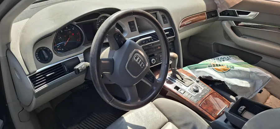 Wnętrze samochodu marki Audi z widocznym kierownicą z logo Audi, zegarami przyrządów, deska rozdzielcza w kolorze beżowym z drewnianymi wstawkami. Na środkowej konsoli znajduje się przyciski sterujące, dźwignia zmiany biegów oraz uszkodzona torba z napisem BINGO i częściowo widocznym napisem TOWER. Wnętrze wykazuje oznaki zużycia, w tym uszkodzenia na konsoli.