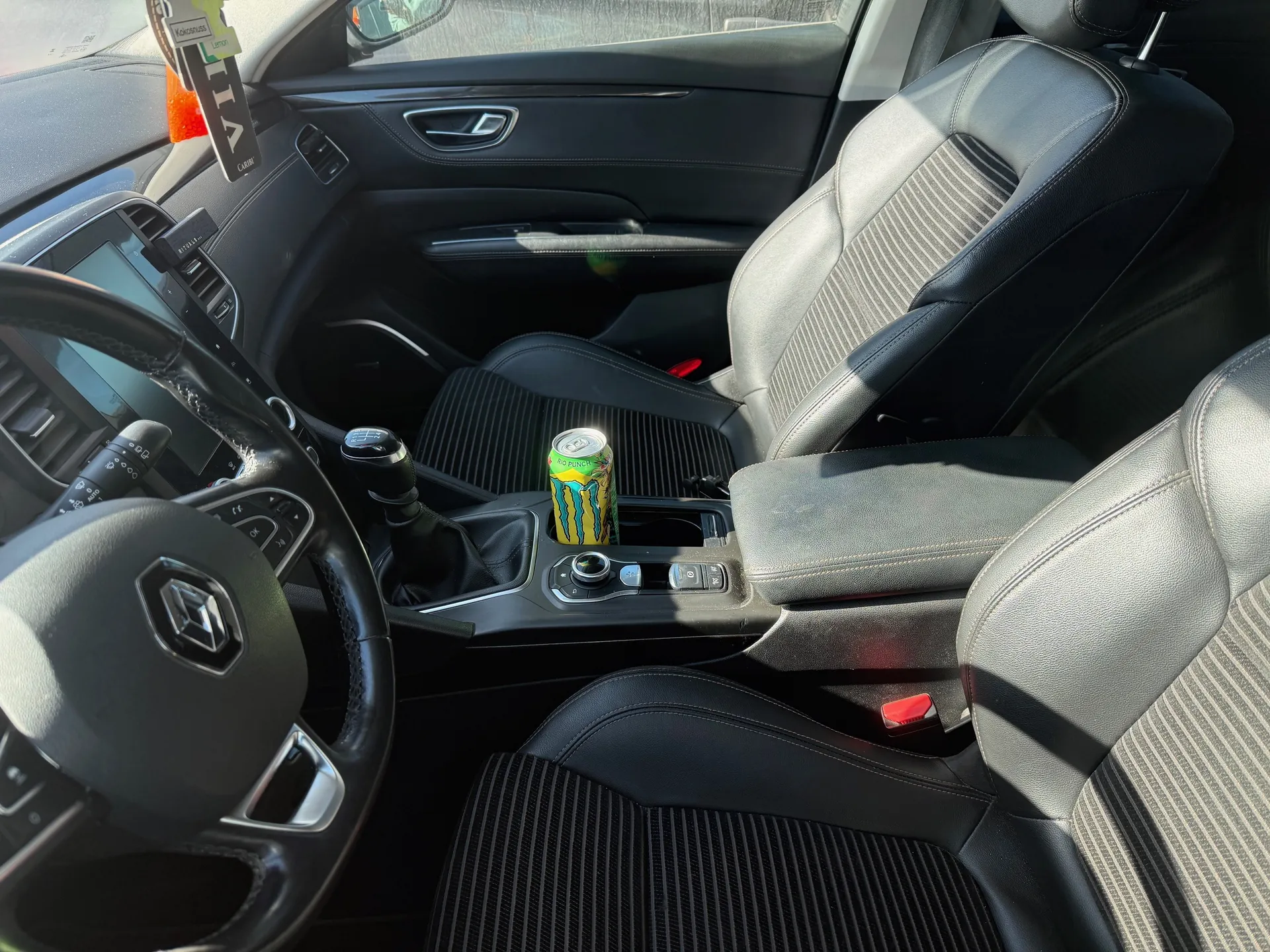 Wnętrze samochodu z czarną skórzaną tapicerką. Widoczne kierownica z logo, dźwignia zmiany biegów, puszka napoju Monster Energy w uchwycie na konsoli środkowej, siedzenia z przeszyciami, ekran na desce rozdzielczej, pomarańczowy przedmiot na desce rozdzielczej oraz wiszący znaczek z napisem "LIV" i "CABIN" na lusterku wstecznym.