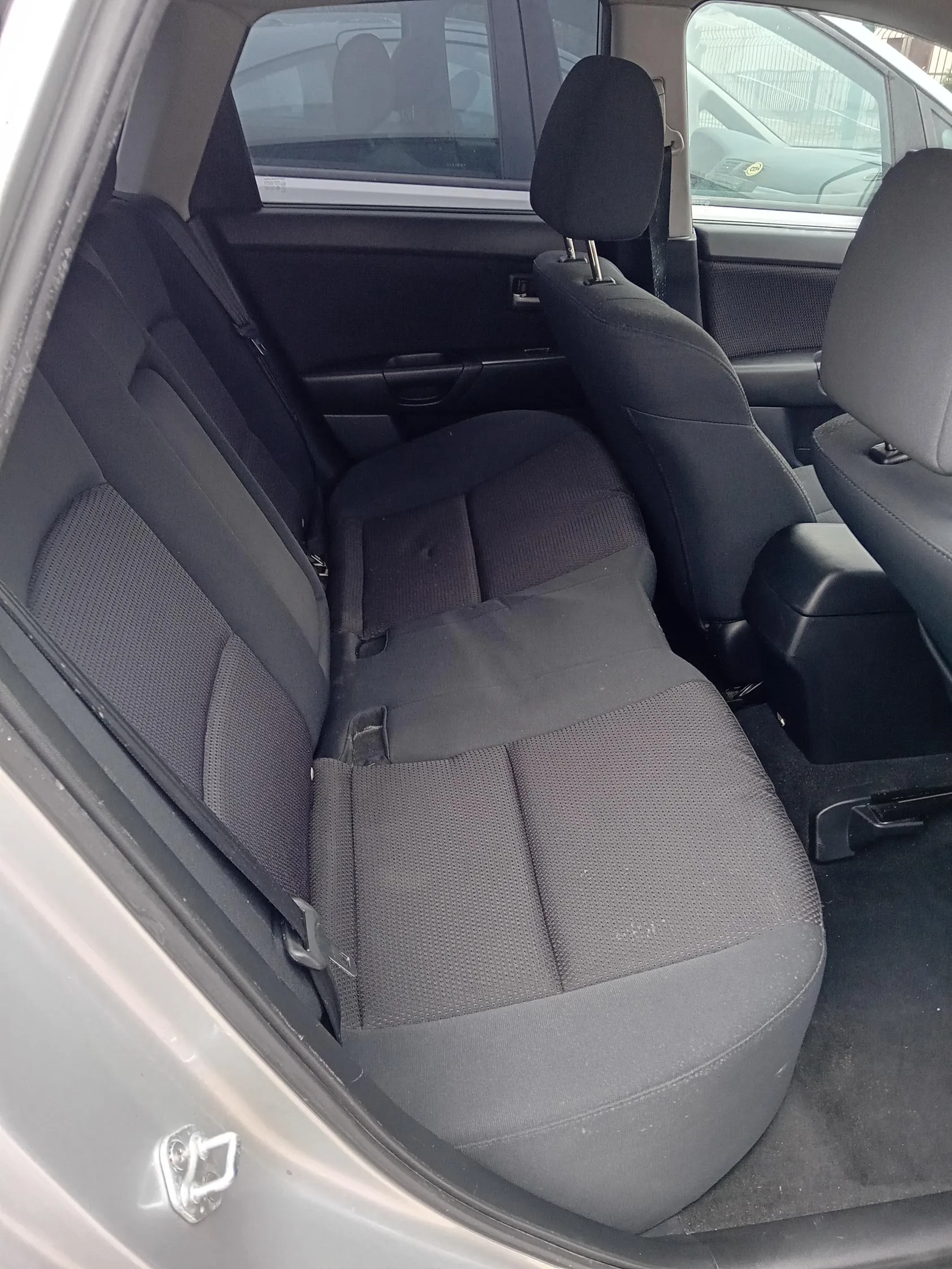 Wnętrze samochodu z widokiem na czarne tkaninowe siedzenia tylnego rzędu. Widoczne są poduszki zagłówkowe, otwarte drzwi z uchwytem i oknem. W tle przez szybę widać inny samochód.