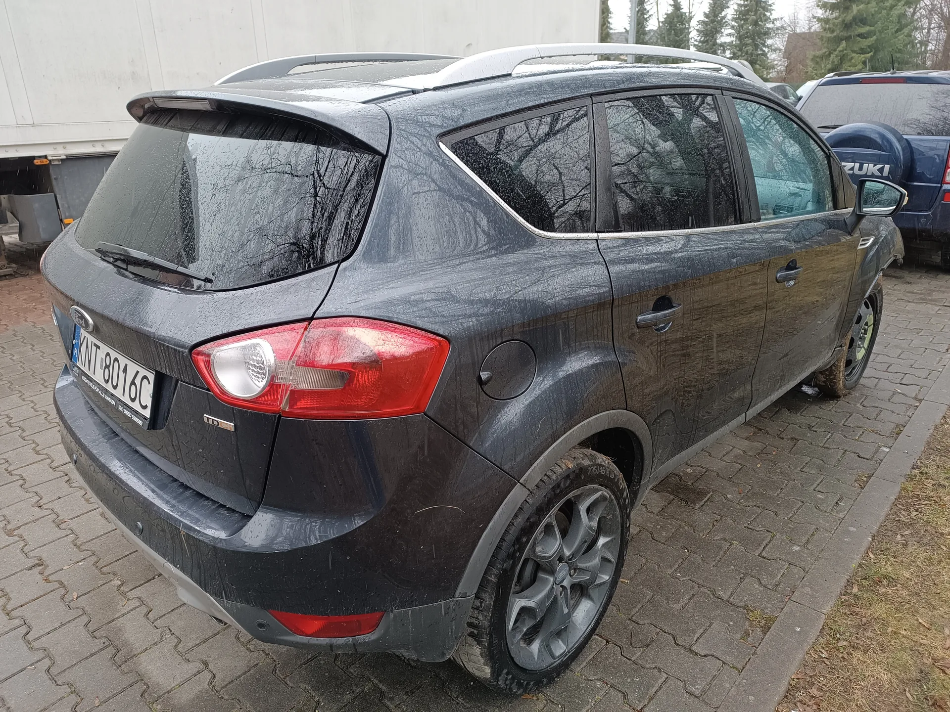 Ciemnoszary SUV marki Ford z widocznym logo na tylnej szybie, zaparkowany na brukowanym placu. Samochód jest zabrudzony błotem i kroplami wody. Widoczna tablica rejestracyjna KNT 8016C oraz czerwone światła tylne. W tle biały naczepa, niebieski samochód Suzuki oraz drzewa. Odbicia drzew na szybach pojazdu. Samochód wyposażony w dachowy bagażnik i srebrne felgi.