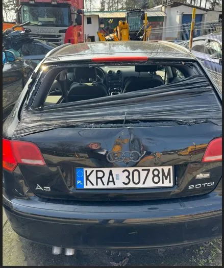 Czarny samochód Audi A3 widziany od tyłu z widoczną tablicą rejestracyjną KRA 3078M z flagą PL. Na bagażniku widoczne uszkodzenie oraz naklejka z nieczytelnym motywem. Po lewej stronie napis A3, po prawej 2.0 TDI. W tle inne pojazdy, czerwony ciężarówka, żółte maszyny budowlane i ogrodzenie. Tekst na tablicy rejestracyjnej jest czytelny i poziomo ustawiony.