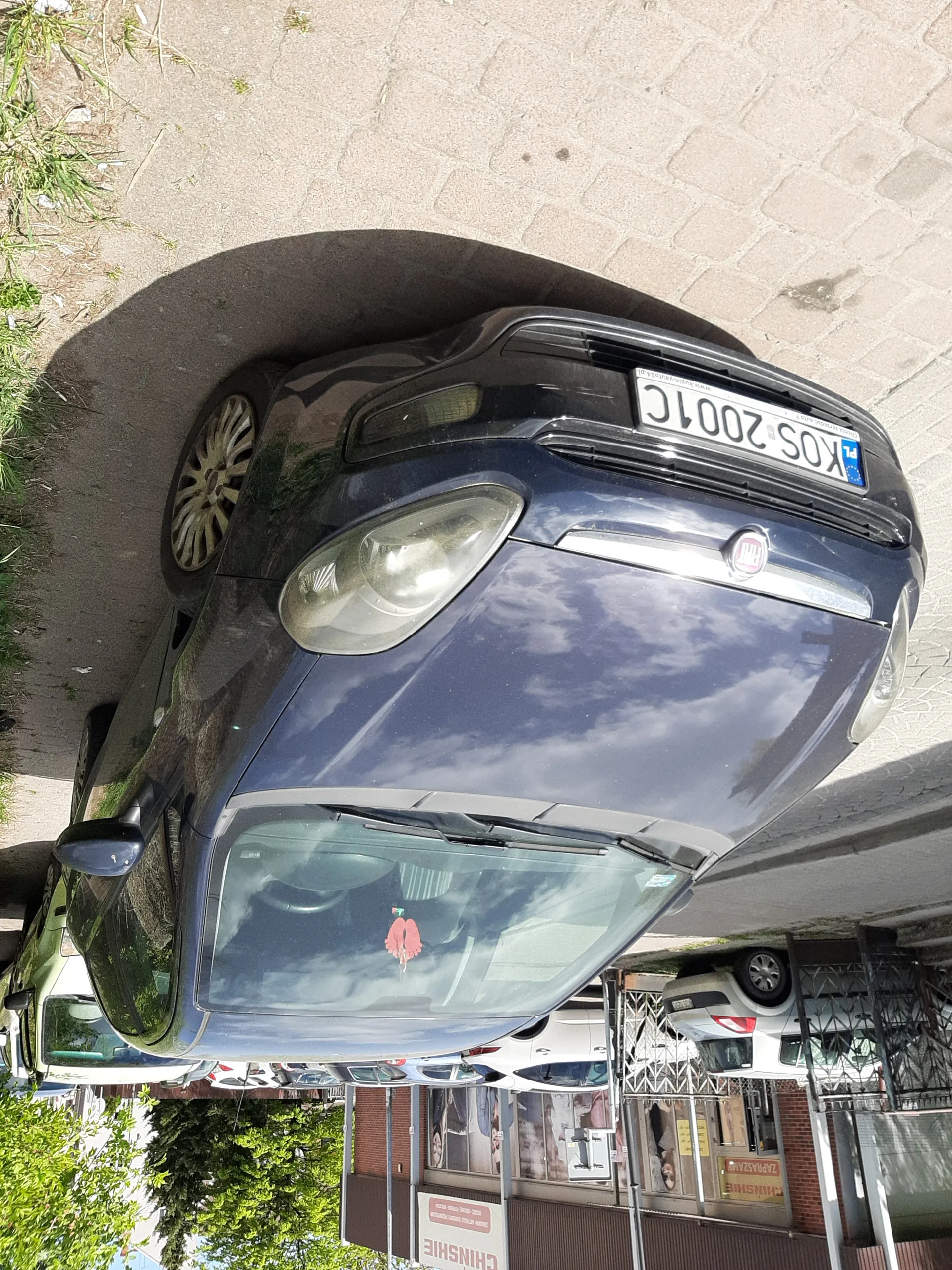 Ciemnoniebieski samochód marki Fiat zaparkowany na brukowanej powierzchni. Widoczna tablica rejestracyjna KOS 2001C z niebieskim pasem po lewej stronie. Na przedniej szybie naklejka w kształcie czerwonej dłoni. W tle budynki z sklepami, w tym tablica z napisem CHINSHE oraz czerwony znak ZAPRASZAMY. Inne zaparkowane samochody, drzewa i chodnik. Słoneczna pogoda, odbicia chmur na karoserii.