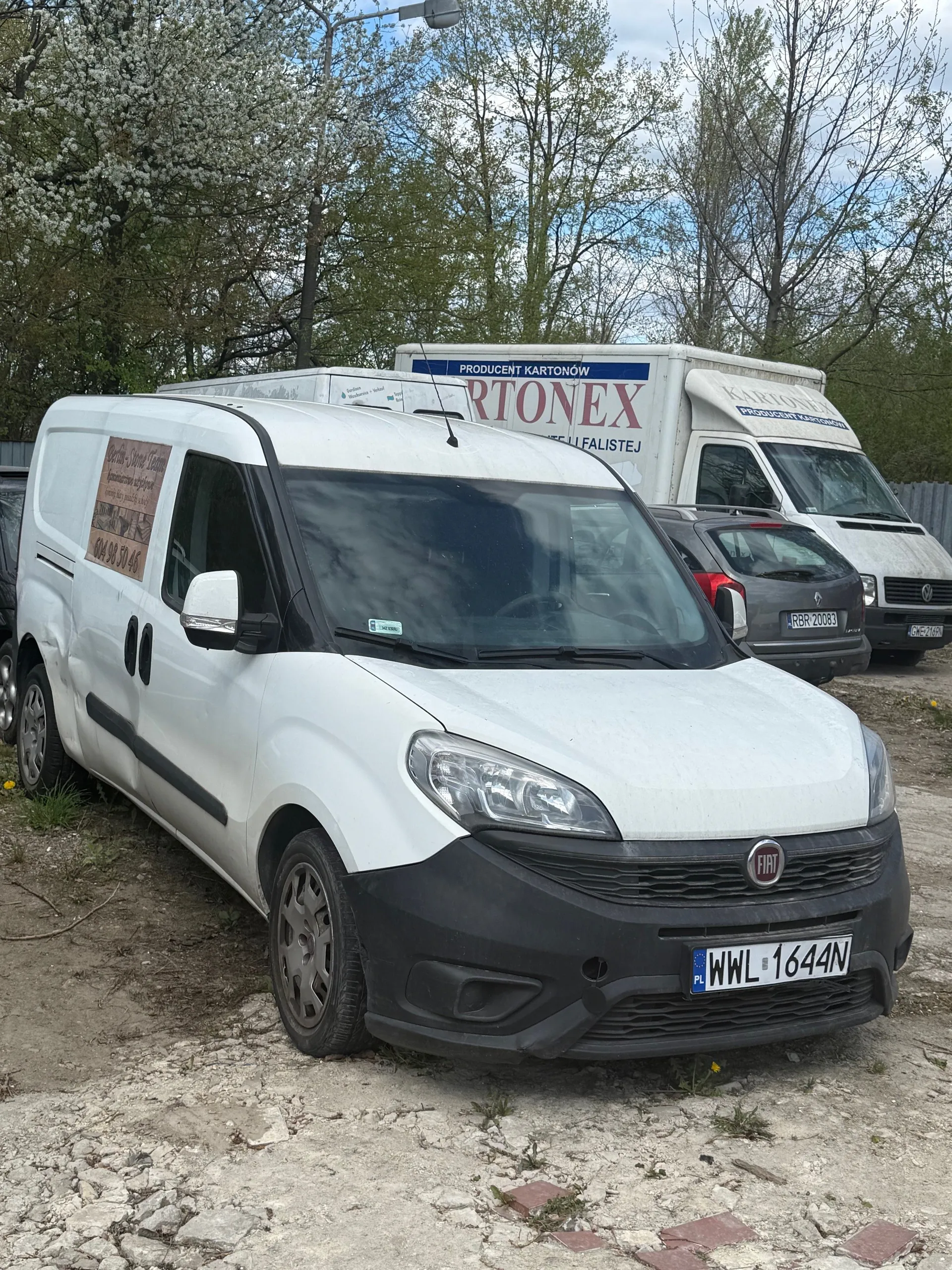 Biały furgon marki Fiat z tablicą rejestracyjną WWL 1644N zaparkowany na pękniętej nawierzchni z widoczną trawą. Na boku furgona znajduje się brązowy plakat z nieczytelnym tekstem. Za nim stoi szary samochód osobowy z tablicą RBR 20083 oraz biały naczepowy samochód dostawczy z napisem "PRODUCENT KARTONÓW" i "KARTONEX" na boku. Tło stanowią drzewa z zielonymi liśćmi i częściowo zachmurzone niebo. Wszystkie pojazdy są ustawione na nierównym terenie z kamieniami i trawą.
