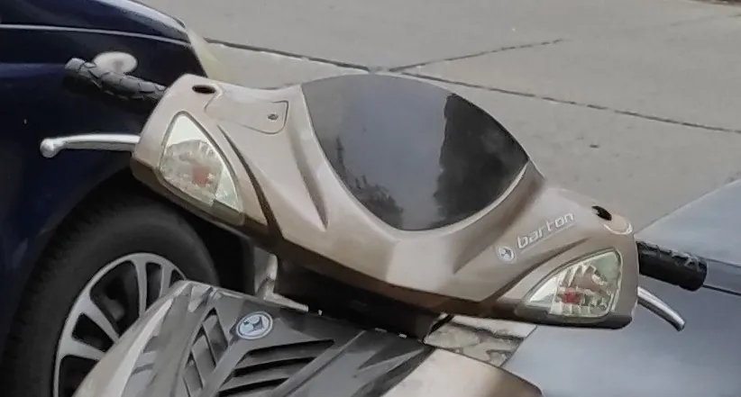 Zbliżenie na kierownicę i przedni panel skuteru w kolorze beżowym. Widoczna jest czarna szyba osłaniająca zegary, dwa kierunkowskazy oraz napis 'barton' po prawej stronie. W tle widoczny jest fragment niebieskiego samochodu i szary chodnik.