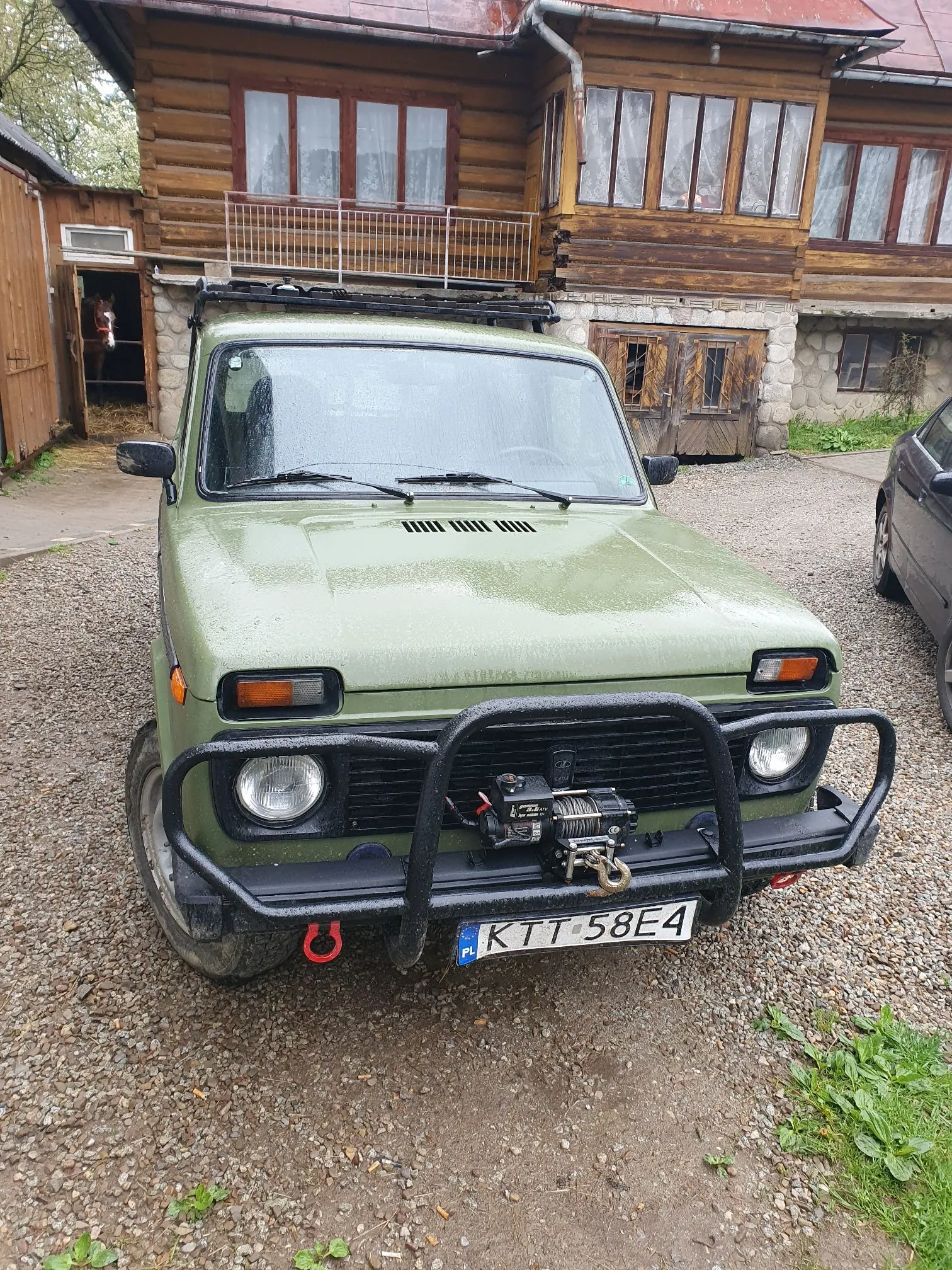 Zielony samochód terenowy marki Lada Niva zaparkowany na żwirowym podjeździe przed drewnianym domem. Pojazd jest wyposażony w czarny bagażnik dachowy, czarną ramę ochronną z przodu (bullbar) z zamontowaną wyciągarką oraz czerwoną haką holowniczą. Tablica rejestracyjna samochodu to KTT 58E4. W tle widoczny jest dwukondygnacyjny budynek z ciemnego drewna z balkonem i oknami.