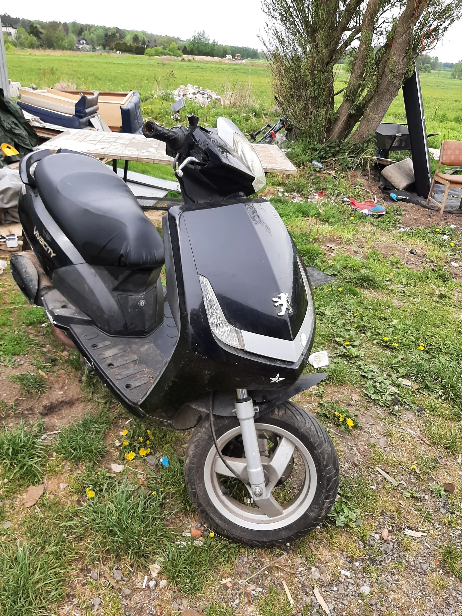 Czarny skuter Peugeot Vivacity stojący na trawie z żółtymi kwiatami. W tle widoczne są różne przedmioty: krzesła, stół, rower oraz rozrzucone materiały. Za nimi rozciągają się zielone pola i drzewa. Trawa jest częściowo zasłana śmieciem i drobnymi przedmiotami.