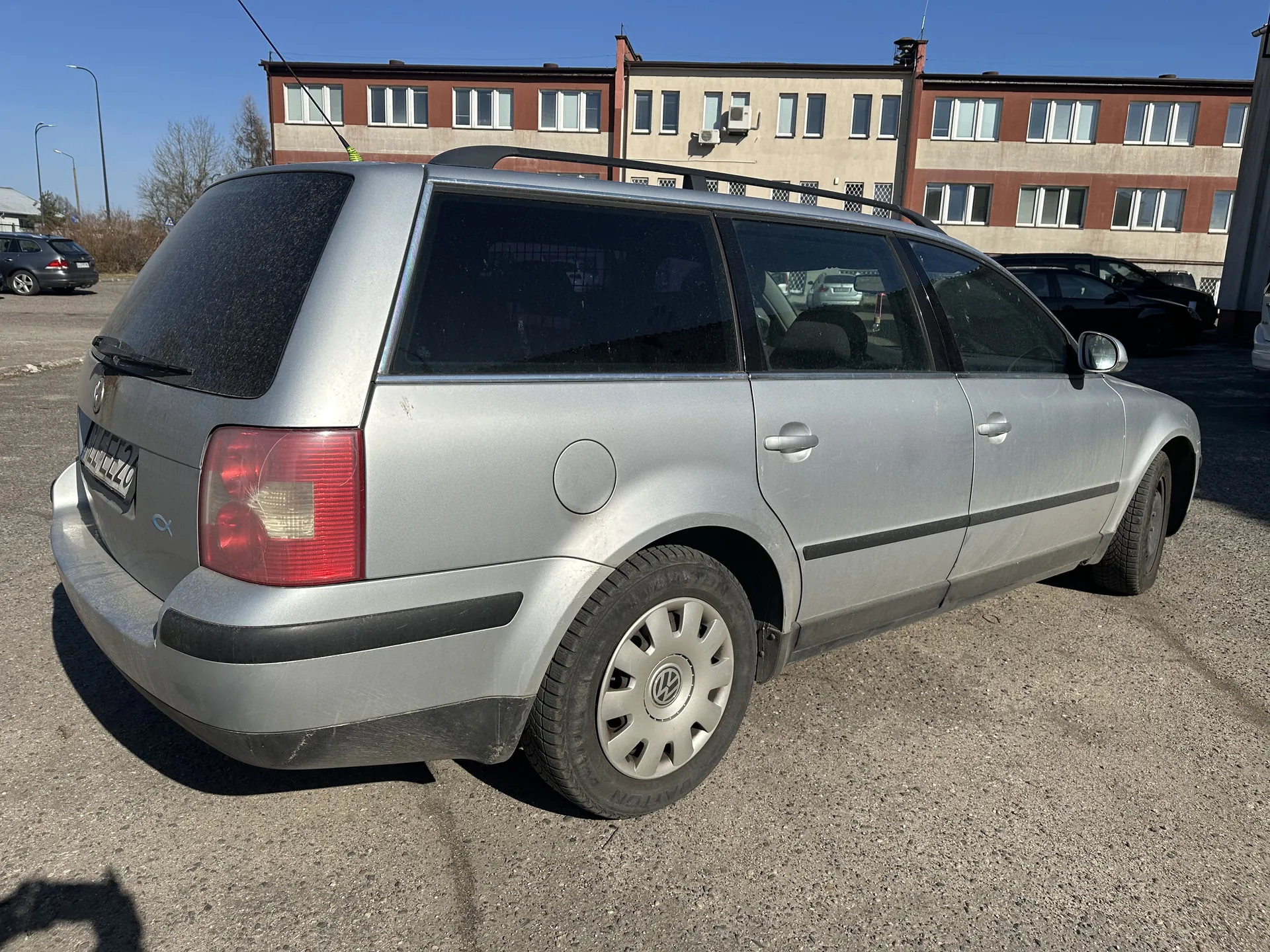 Srebrny samochód typu kombi marki Volkswagen zaparkowany na asfaltowym placu parkingowym. Widok od tyłu i boku. Na tylnej szybie widoczna wycieraczka, czerwone światła tylnego oświetlenia oraz tablica rejestracyjna z numerem H47220. Samochód wyposażony w dachowy bagażnik. W tle budynki o czerwono-beżowej elewacji z wieloma oknami, inne zaparkowane pojazdy oraz jasne niebo bez chmur.