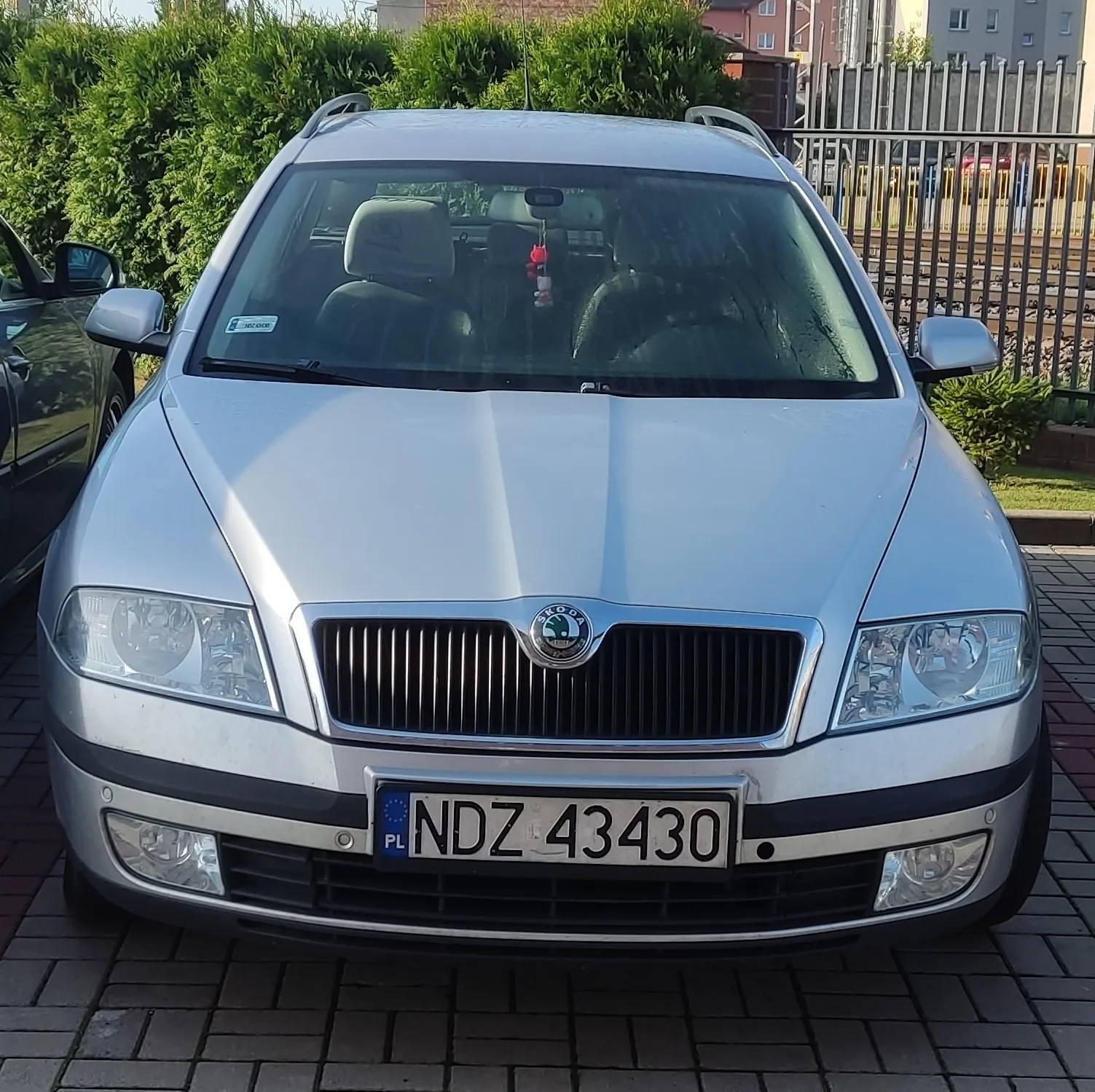 Srebrny samochód marki Skoda zaparkowany przodem do widza na kostce brukowej. Pojazd posiada polską tablicę rejestracyjną o numerze NDZ 43430. W tle widoczne są zielone krzewy oraz metalowy płot.