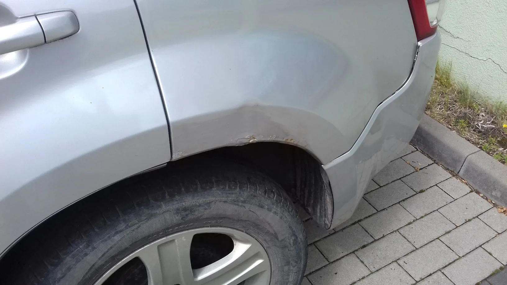 Zbliżenie na tylny błotnik srebrnego samochodu. Nad nadkolem widoczne jest wgniecenie oraz niewielkie ogniska korozji i odpryski lakieru. W dolnej części obrazu widoczna jest opona i fragment felgi aluminiowej, a samochód zaparkowany jest obok krawężnika i chodnika z szarej kostki brukowej.