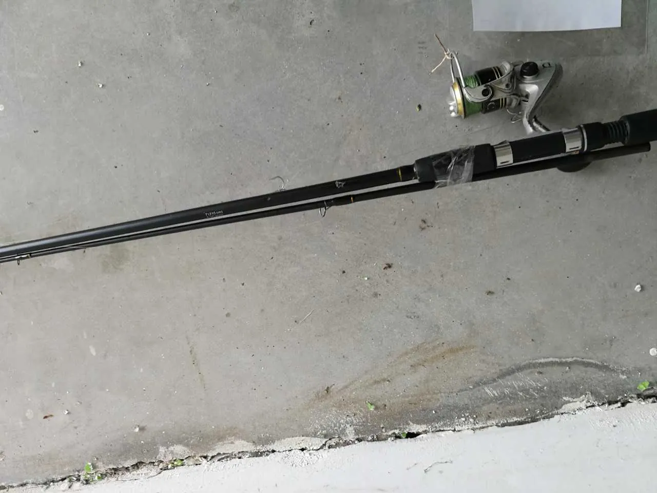 Czarny wędkarski kij z zielono-srebrną podajnikową zębatką leżący na szarym betonowym podłożu. W tle widoczne są drobne śmieci, pęknięcie w betonie oraz biały arkusz papieru w prawym górnym rogu. Na kiju znajduje się niewielki, nieczytelny napis.