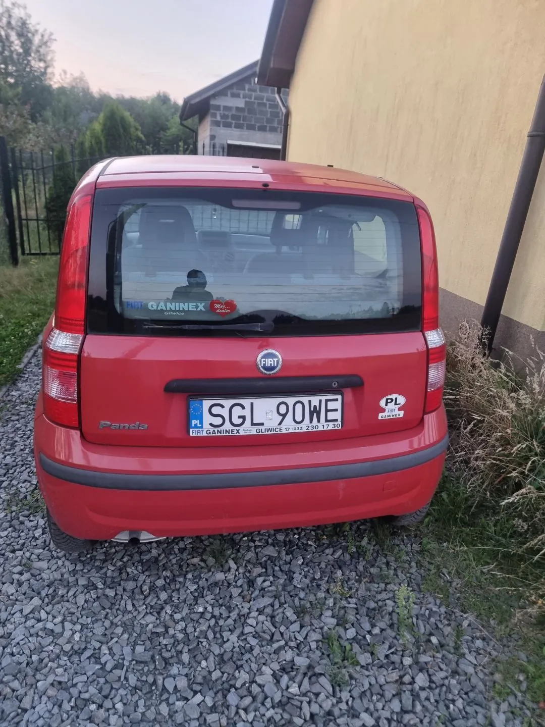 Tylny widok czerwonego samochodu marki Fiat Panda zaparkowanego na żwirowym podjeździe obok żółtej ściany budynku. Samochód ma polską tablicę rejestracyjną o numerze SGL 90WE. Na tylnej szybie znajduje się naklejka z napisem 'GANINEX Gliwice', a pod tablicą rejestracyjną widnieje napis 'FIAT GANINEX GLIWICE 17 ul. Suli 230 17 30'. Po prawej stronie tablicy znajduje się mała naklejka z napisem 'PL FIAT GANINEX'.