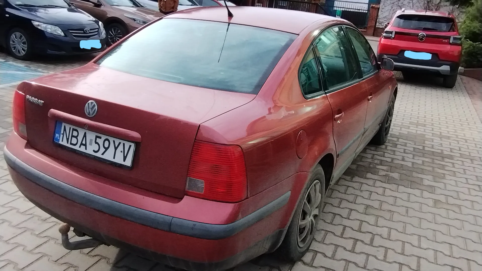 Czerwony samochód Volkswagen Passat z widocznym tyłem, tablicą rejestracyjną PL NBA 59YV oraz napisem PASSAT po lewej stronie bagażnika. W tle czarny samochód marki Toyota, czerwony SUV oraz brukowana powierzchnia parkingu. Widoczne są także elementy otoczenia: ogrodzenie, budynki i drzewa.