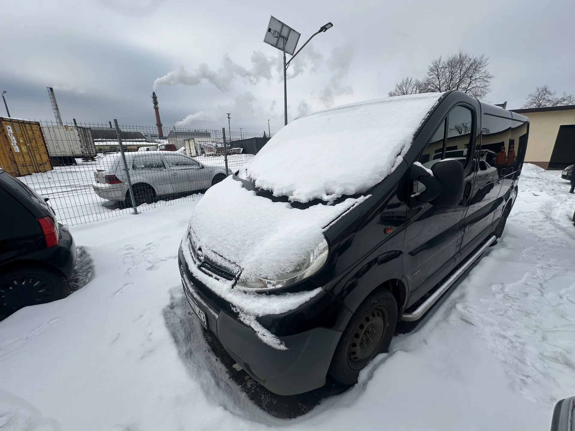 Czarny samochód dostawczy pokryty śniegiem na pierwszym planie, zaparkowany na śnieżnej powierzchni. W tle widoczne są inne pojazdy (srebrny i czarny samochód), metalowe ogrodzenie, przemysłowe budynki, komin z dymem, latarnia uliczna z panelem słonecznym oraz pozbawione liści drzewa. Niebo jest zachmurzone. Śnieg pokrywa całą scenę, w tym dach i przednią część dostawczego pojazdu.