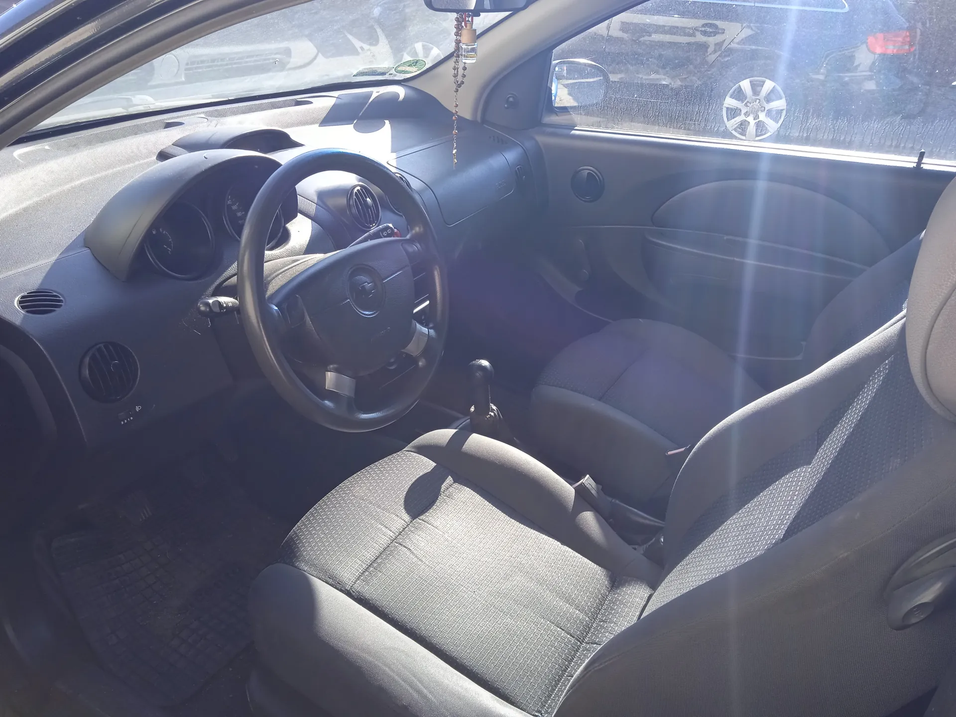 Wnętrze samochodu z widoczną kierownicą po lewej stronie, tablicą przyrządów z trzema okrągłymi wskaźnikami, szarymi siedzeniami z tkaniną w drobny wzór, dźwignią zmiany biegów oraz przednią szybą pokazującą zaparkowane samochody na zewnątrz. W lusterku wstecznym widoczny wiszący ozdobny element. Światło słoneczne wpada przez okna, tworząc refleksy.