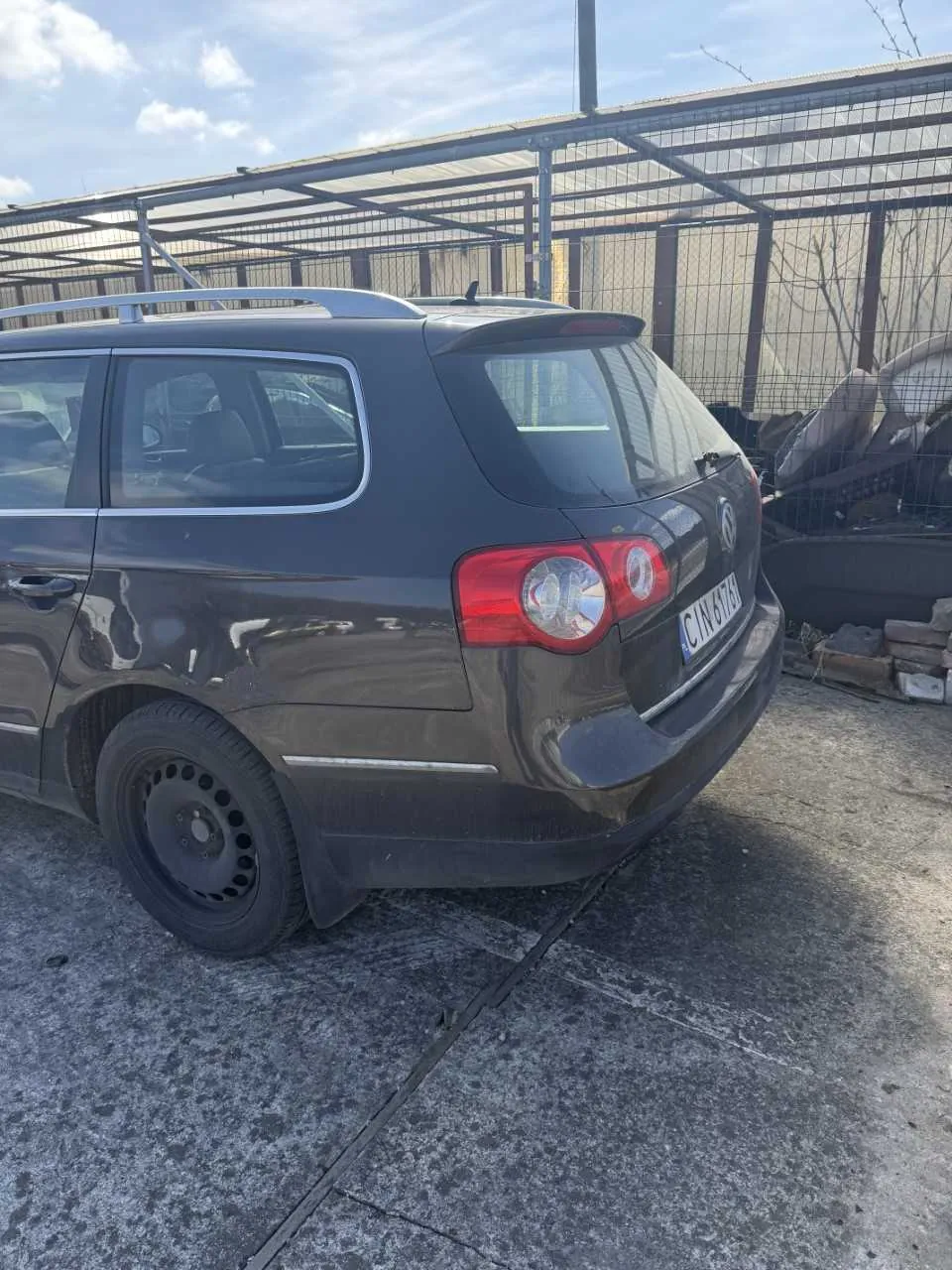 Ciemnoszary samochód typu kombi zaparkowany na betonowej powierzchni. Widok z tyłu i boku. Tablica rejestracyjna z napisem CIN 678. Na dachu dachówka. W tle metalowe ogrodzenie, przemysłowa konstrukcja z dachem oraz stosy starych części samochodowych. Niebo częściowo zachmurzone.