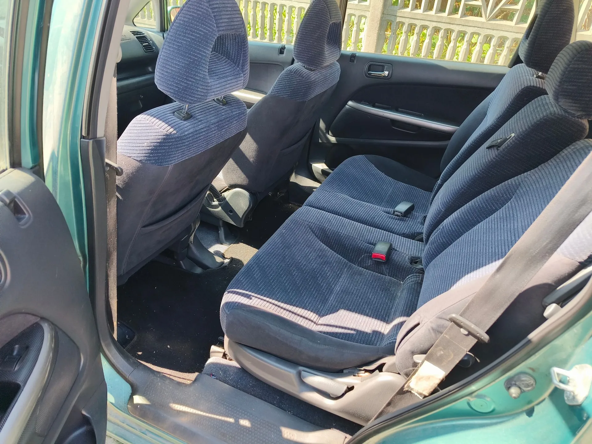 Wnętrze samochodu z widokiem na tylną kanapę i przednie fotele. Tapicerka siedzeń jest ciemnoniebieska w prążki. Widoczne są pasy bezpieczeństwa oraz fragmenty drzwi i nadwozia w kolorze zielonym.