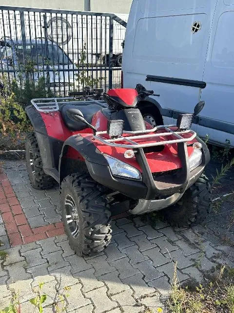 Czerwony quad marki Honda zaparkowany na kostce brukowej obok białego vana. Pojazd posiada srebrny bagażnik z przodu i z tyłu, duże terenowe opony oraz dodatkowe lampy LED zamontowane na przednim stelażu.