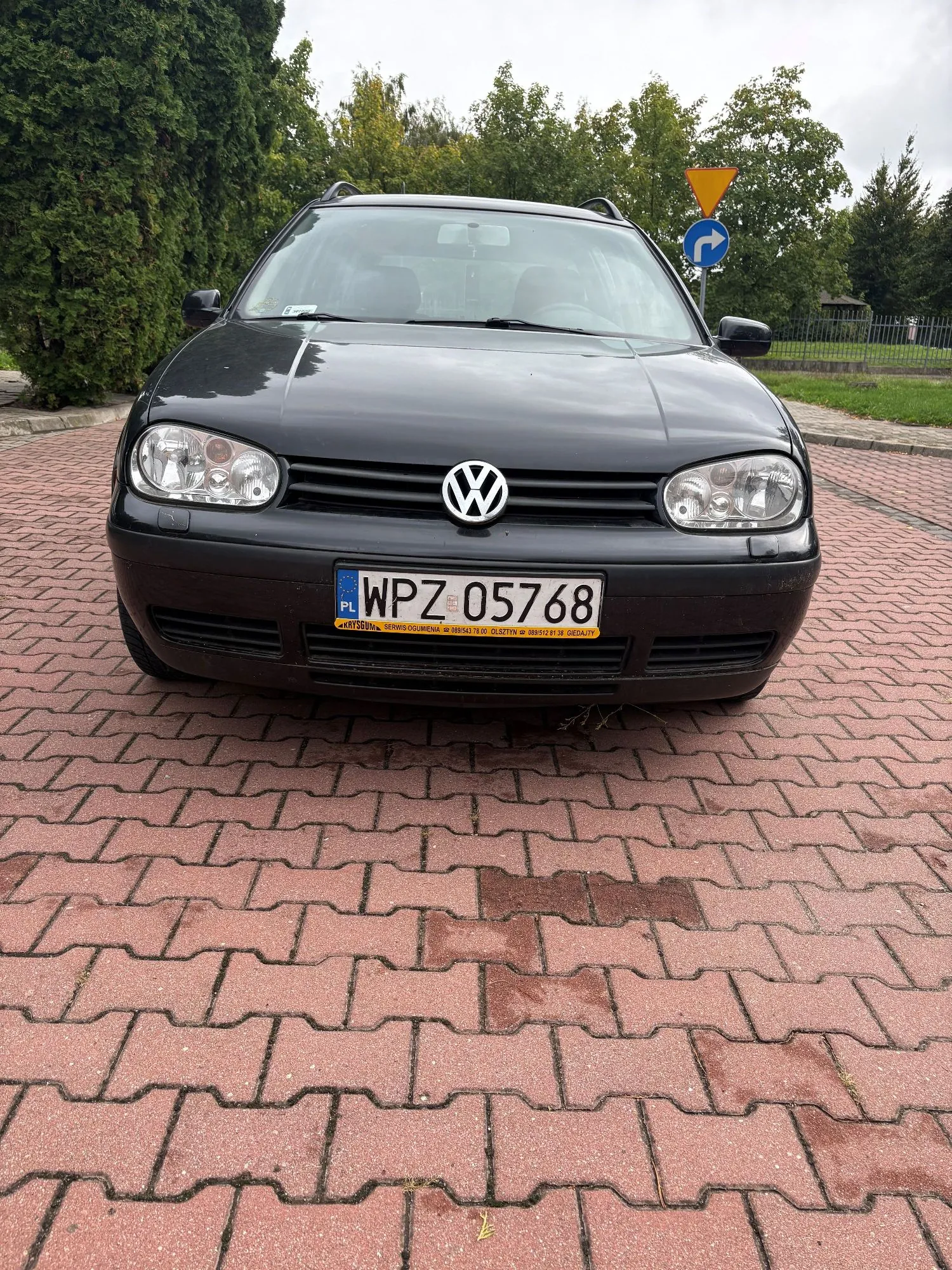 Czarny samochód marki Volkswagen zaparkowany przodem do widza na czerwonej kostce brukowej. Na samochodzie widoczna jest tablica rejestracyjna o numerze WPZ 05768. Poniżej tablicy znajduje się żółty pasek z tekstem: 'SERWIS COLUMNA sp. z o.o. 58352 76 00 GDAŃSK tel. 58 552 81 81 GDAŃSK'. W tle widać zielone drzewa oraz znak drogowy z niebieską strzałką i żółtym trójkątem.