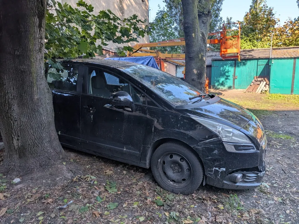 Czarny, zakurzony samochód osobowy typu minivan zaparkowany blisko pnia dużego drzewa. Pojazd jest uszkodzony w przedniej części po prawej stronie, gdzie widać wgniecenia zderzaka i maski. W tle widoczny jest pomarańczowy podnośnik koszowy, zielone drzwi garażowe oraz liściaste drzewa.