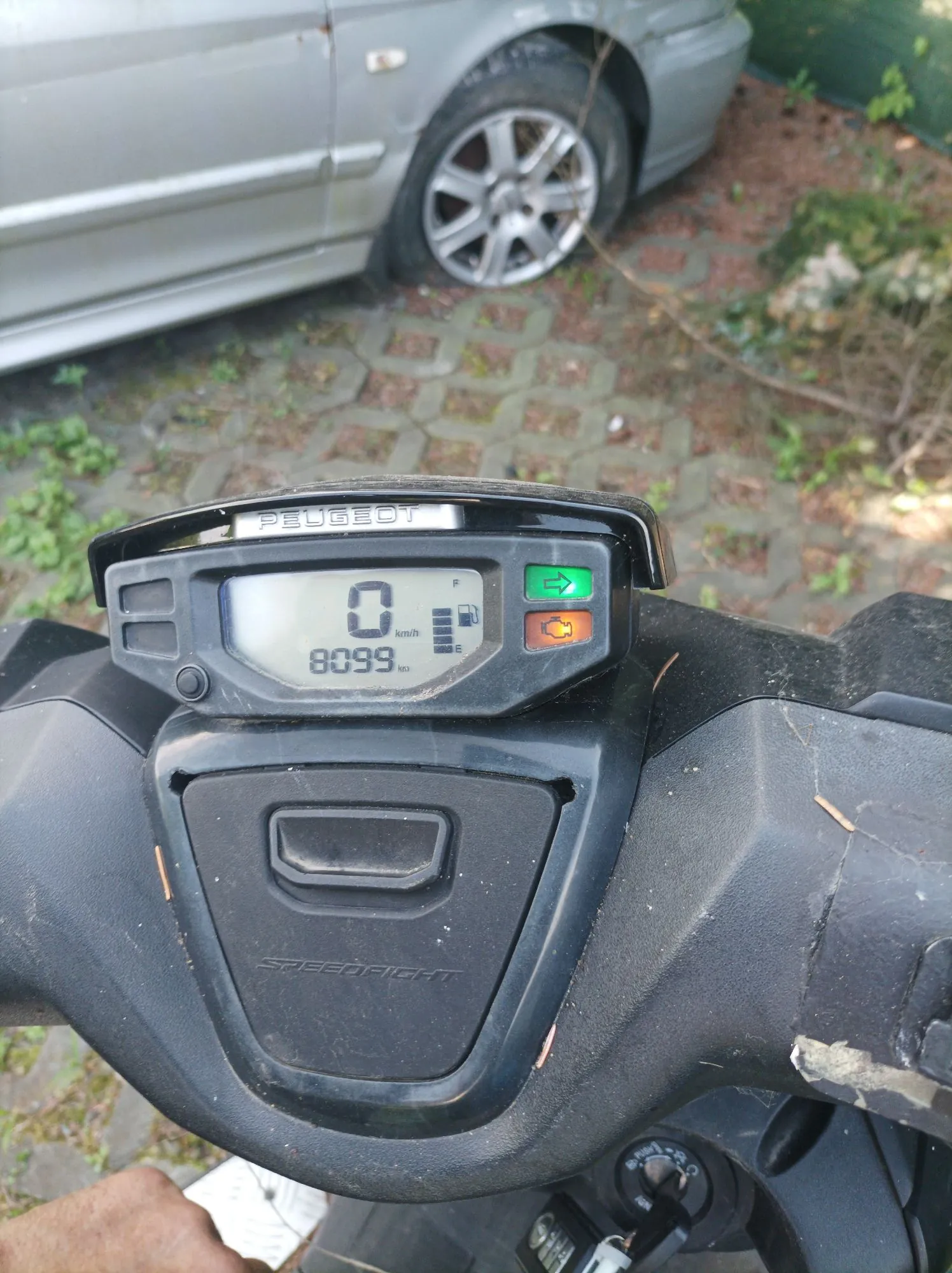 Zbliżenie na deskę rozdzielczą czarnego skutera marki Peugeot. Cyfrowy wyświetlacz pokazuje prędkość 0 km/h oraz przebieg 8099 km. Po prawej stronie wyświetlacza znajduje się zielona strzałka kierunkowskazu oraz pomarańczowa ikona kontrolna. Poniżej wyświetlacza widnieje napis SPEEDFIGHT.