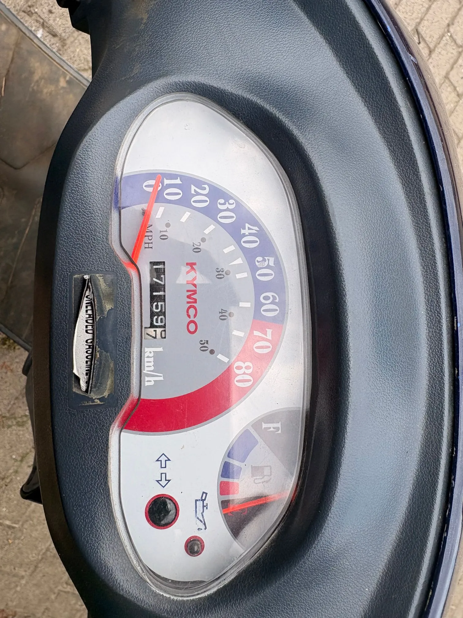 Zbliżenie na licznik prędkości skutera marki KYMCO. Prędkościomierz posiada skalę w km/h od 0 do 80 oraz mniejszą skalę w MPH. Wskazówka prędkości znajduje się na poziomie 0. Widoczny jest licznik przebiegu z wartością 17159.7 km/h. Poniżej znajduje się wskaźnik poziomu paliwa z oznaczeniami F i R oraz czerwona kontrolka. Całość jest osadzona w czarnej, plastikowej obudowie.