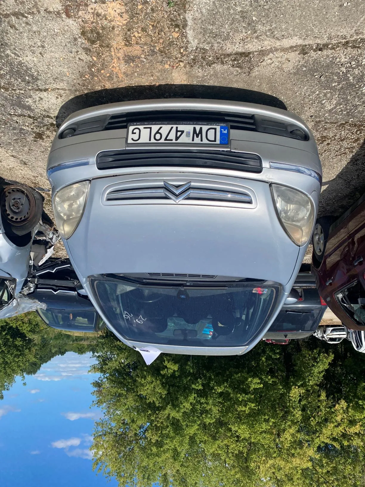 Srebrny samochód osobowy marki Citroën z polską tablicą rejestracyjną DW 479LG, zaparkowany na betonowym placu. Pojazd jest widoczny z przodu, w tle znajdują się inne uszkodzone samochody oraz zielone drzewa pod niebieskim niebem z białymi chmurami.