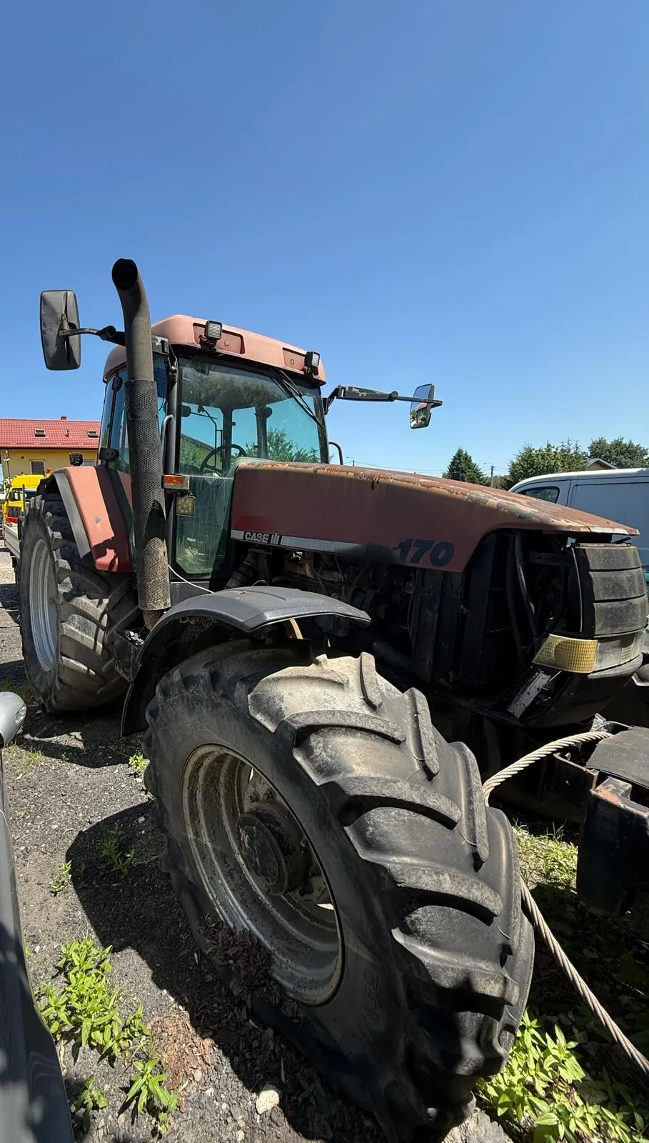 Czerwony traktor marki Case IH model 170, zaparkowany na żwirowym podłożu. Widoczny jest z perspektywy z przodu i z boku, z dużymi, błotnistymi oponami z wyraźnym bieżnikiem. Na masce widnieje napis 'CASE IH' oraz liczba '170'. W tle widać fragmenty budynków oraz bezchmurne, niebieskie niebo.