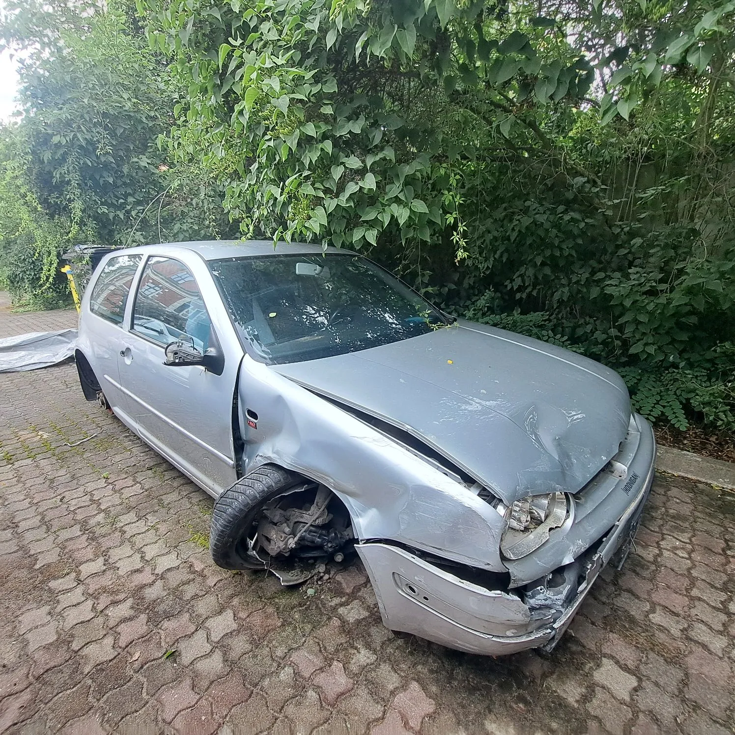 Srebrny samochód osobowy marki Volkswagen Golf z poważnymi uszkodzeniami przodu. Przedni prawy błotnik jest wgnieciony, reflektor rozbity, a przedni zderzak częściowo oderwany i pęknięty. Przednie prawe koło jest wykręcone i leży pod kątem względem nadwozia. Auto zaparkowane jest na kostce brukowej na tle gęstych, zielonych krzewów i drzew.