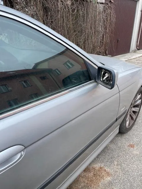 Srebrny samochód widziany z boku, z widocznym lusterkiem bocznym i drzwiami. W tle budynki z brązowymi drzwiami garażowymi oraz suche pnącza na ścianie. W szybie odbija się czerwony budynek z oknami. Samochód stoi na nawierzchni z drobnych kamieni.