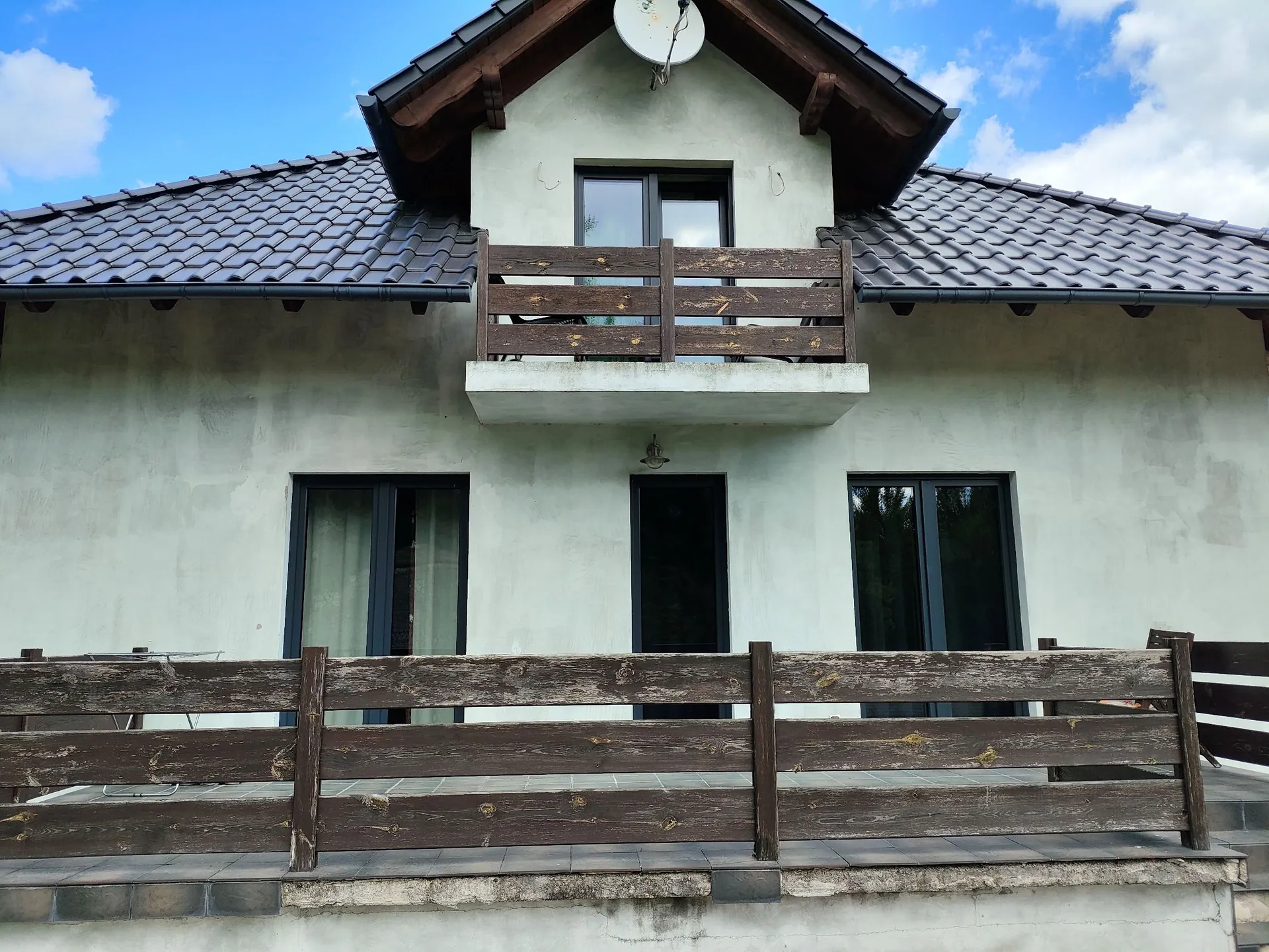 Frontowa elewacja jasnoszarego, dwukondygnacyjnego domu z ciemnym, spadzistym dachem z dachówki. Na piętrze znajduje się mały balkon z drewnianą balustradą i ciemnymi oknami. Na dachu zamontowano białą antenę satelitarną. Na parterze widoczne są dwa duże, ciemne okna oraz ciemne drzwi wejściowe. Przed domem znajduje się szeroki taras z ciemnobrązową, drewnianą balustradą.