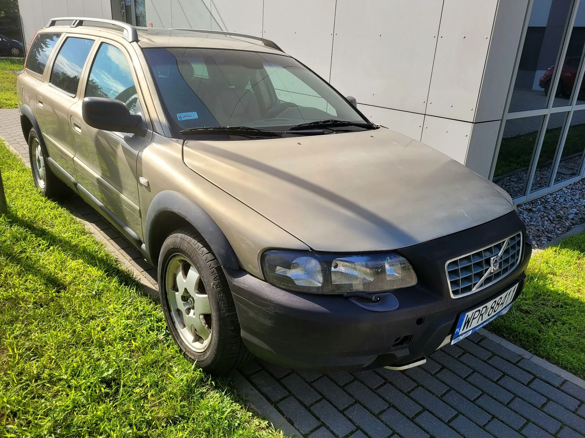 Złoty samochód marki Volvo w nadwoziu kombi, zaparkowany na kostce brukowej obok trawnika i nowoczesnego białego budynku. Pojazd ma czarne plastikowe nadkola i zderzak, a na tablicy rejestracyjnej widnieje napis WPR 88411.