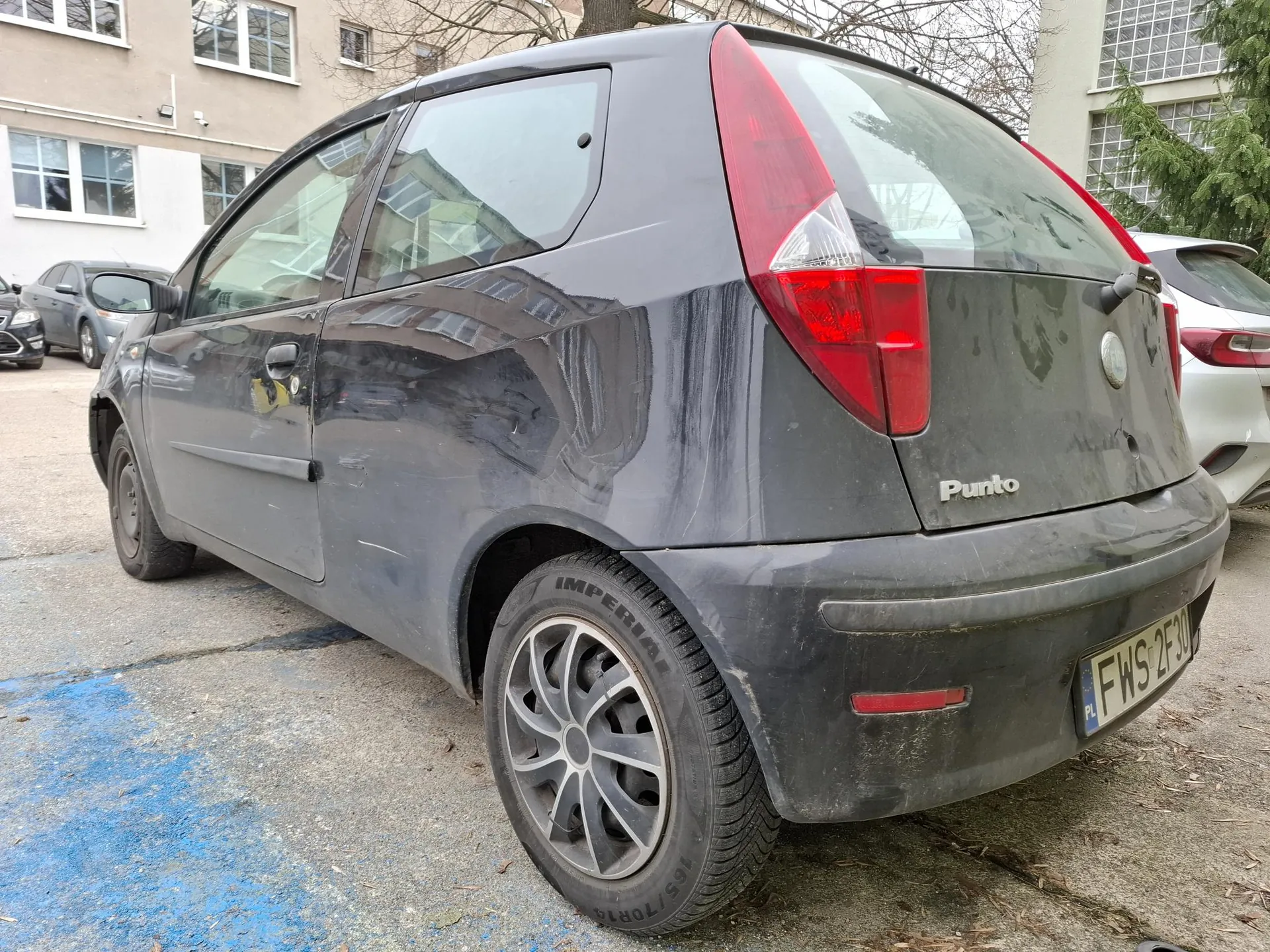 Ciemnoszary samochód Fiat Punto zaparkowany na placu parkingowym. Widoczny tył pojazdu z czerwonymi światłami, tablicą rejestracyjną FWS 2F30 (PL) oraz napisem 'Punto' na bagażniku. Opona z napisem 'IMPERIAL'. W tle budynki z oknami, inne zaparkowane samochody i drzewa. Na asfalcie niebieskie znakowanie.
