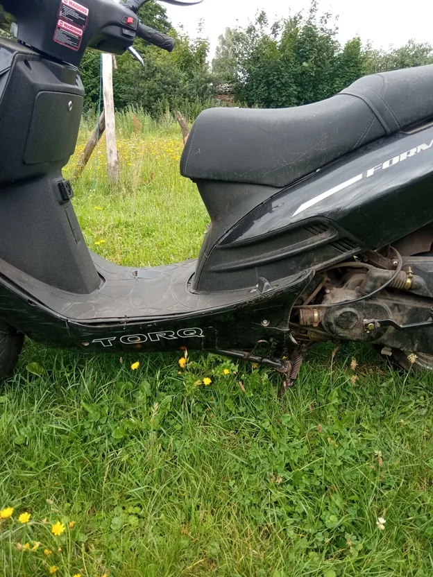 Czarny skuter marki Torq zaparkowany na zielonej trawie z żółtymi kwiatami. Widoczny jest profil pojazdu z siedzeniem, podnóżkiem oraz napisem 'TORQ' na dolnej części nadwozia i fragmentem napisu 'FORM' na boku. W tle znajdują się zielone krzewy i drzewa.