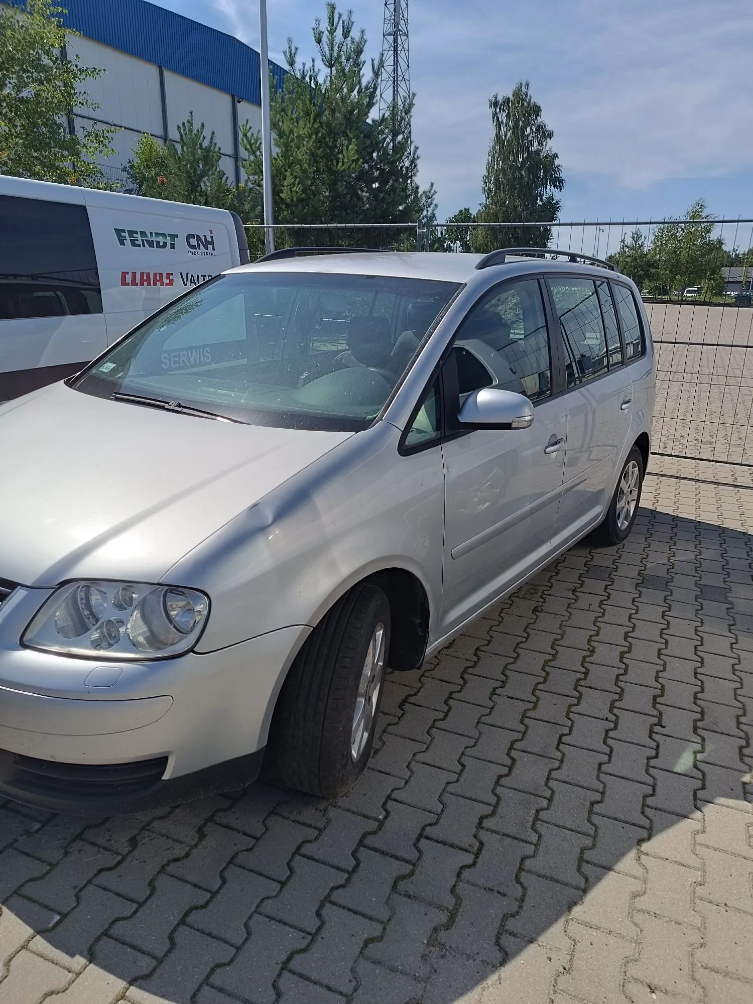 Srebrny samochód typu kombi zaparkowany na kostce brukowej. W tle widoczny jest biały samochód dostawczy z napisami 'FENDT CNH INDUSTRIAL', 'CLAAS VALTRA' oraz 'SERWIS'. W oddali znajdują się zielone drzewa i fragment niebieskiego budynku przemysłowego pod jasnym niebem.