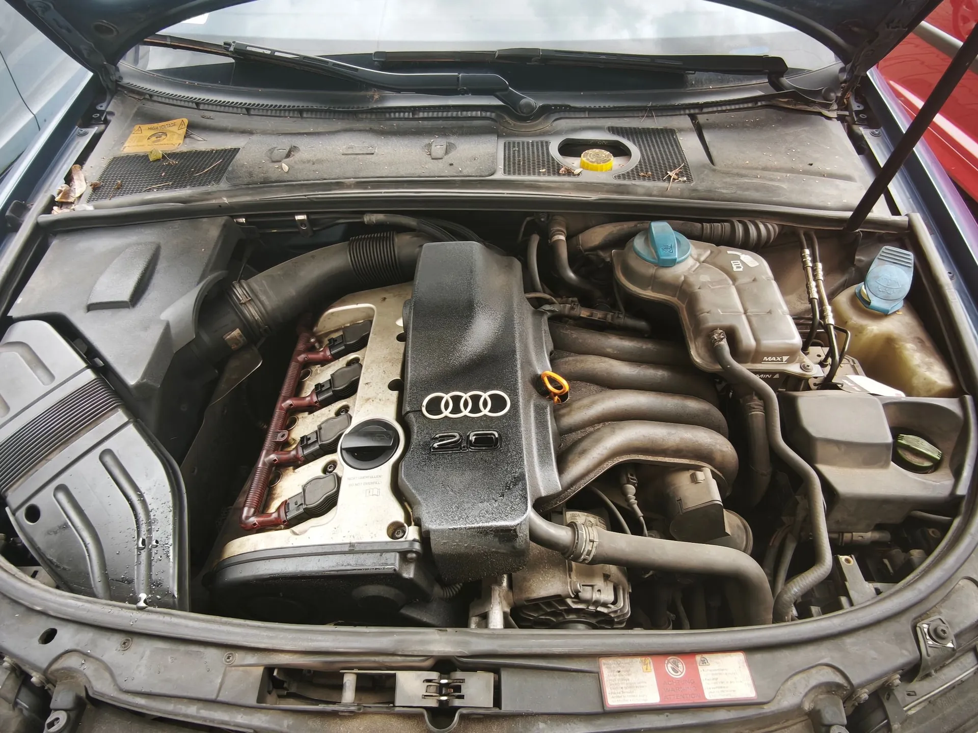 Widok z góry na komorę silnika samochodu marki Audi. W centrum znajduje się silnik z czarną plastikową osłoną z logo Audi i oznaczeniem '2.0'. Po prawej stronie widoczne są zbiorniki na płyny eksploatacyjne z niebieskimi zakrętkami oraz układ rur i przewodów. Silnik i otaczające go elementy są zakurzone.