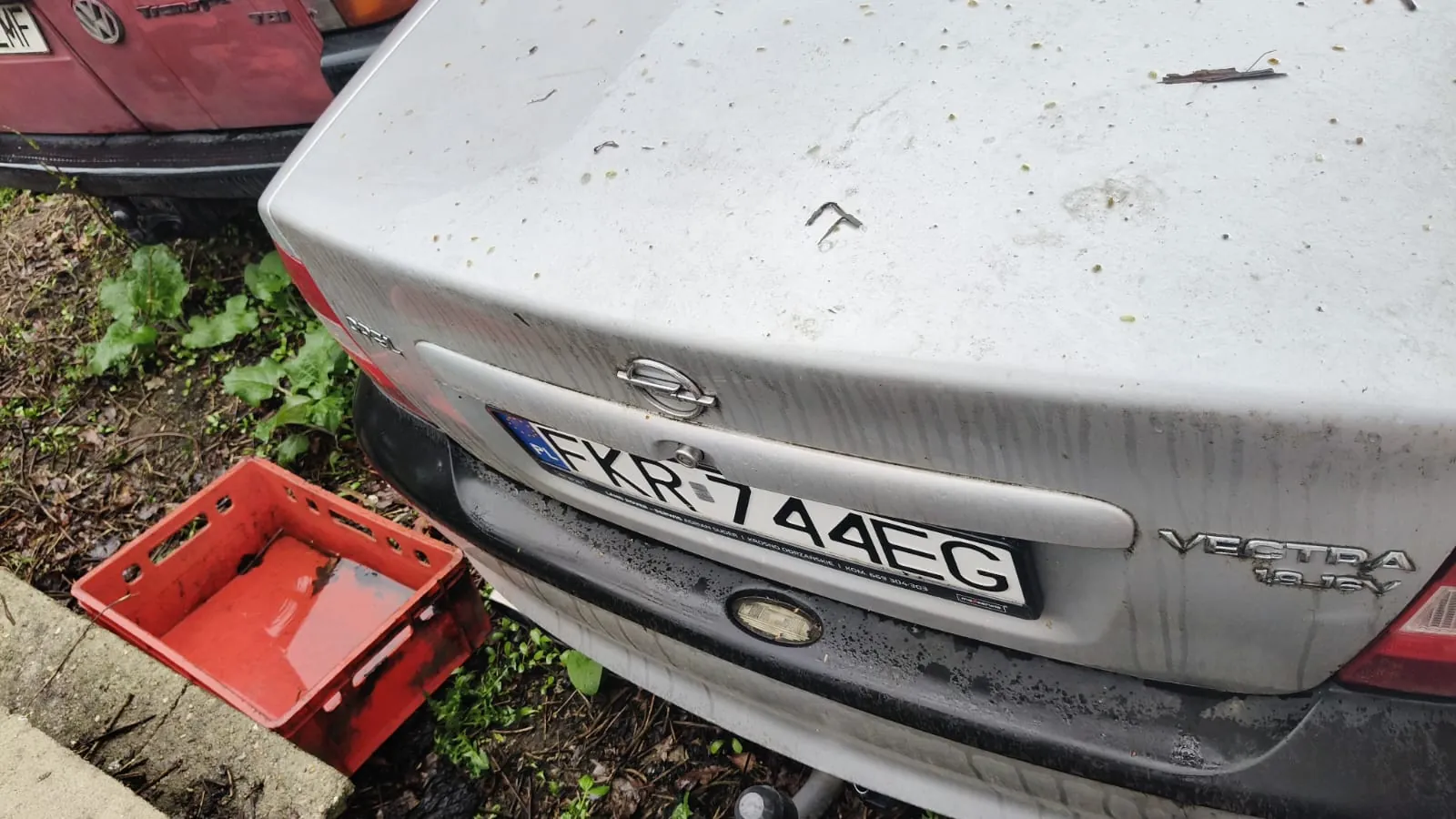 Zbliżenie na tył srebrnego samochodu Opel Vectra z tablicą rejestracyjną FKR 744EG. Na bagażniku widoczne są zabrudzenia i ślady brudu. Po lewej stronie obrazu czerwony plastikowy kosz na ziemi, zielone rośliny oraz fragment czerwonego samochodu w tle. Na bagażniku widoczny napis 'VECTRA 1.8 16V'.