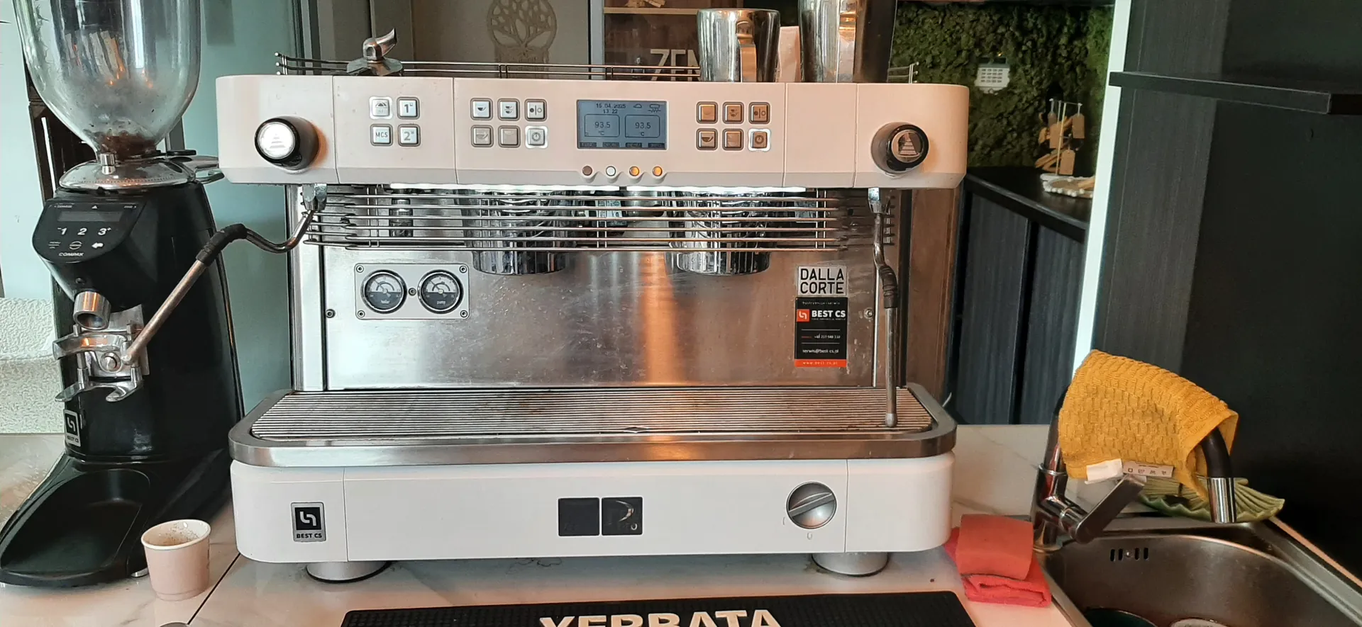 Biała i stalowa maszyna do kawy marki DALLA CORTE z cyfrowym wyświetlaczem, dwoma manometrami i przyciskami. Po lewej stronie czarny zmielacz kawy z przezroczystym zbiornikiem, obok niego mały papierowy kubek. Po prawej zlewozmywak z żółtą ściereczką na kranie i czerwoną ściereczką na blacie. W tle zielona ściana z mchem i półki z metalowymi kubkami. Na maszynie widoczne naklejki z napisami DALLA CORTE, BEST CS oraz cyfrowy wyświetlacz z wartościami 93.5 i 93.5. Na czarnej podkładce przed maszyną widoczny fragment napisu YERRATA.