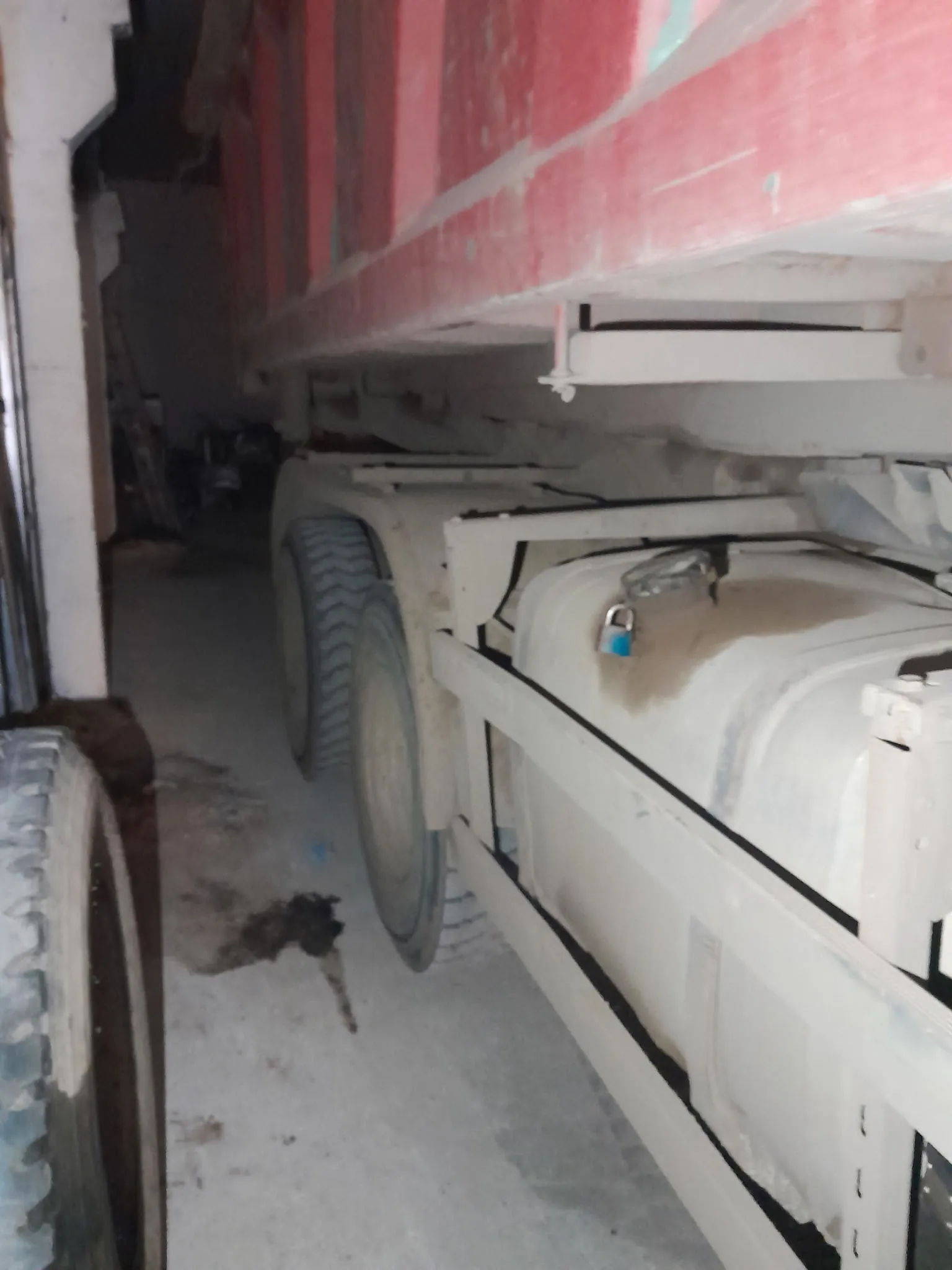 Boczny widok dużego pojazdu ciężarowego z czerwono-białym zbiornikiem. Widoczne są duże opony, biały zbiornik paliwa z niebieskim kłódkiem oraz betonowa podłoga z plamami i śmieciem. Tło jest ciemne, prawdopodobnie garaż lub magazyn. Brak czytelnego tekstu.