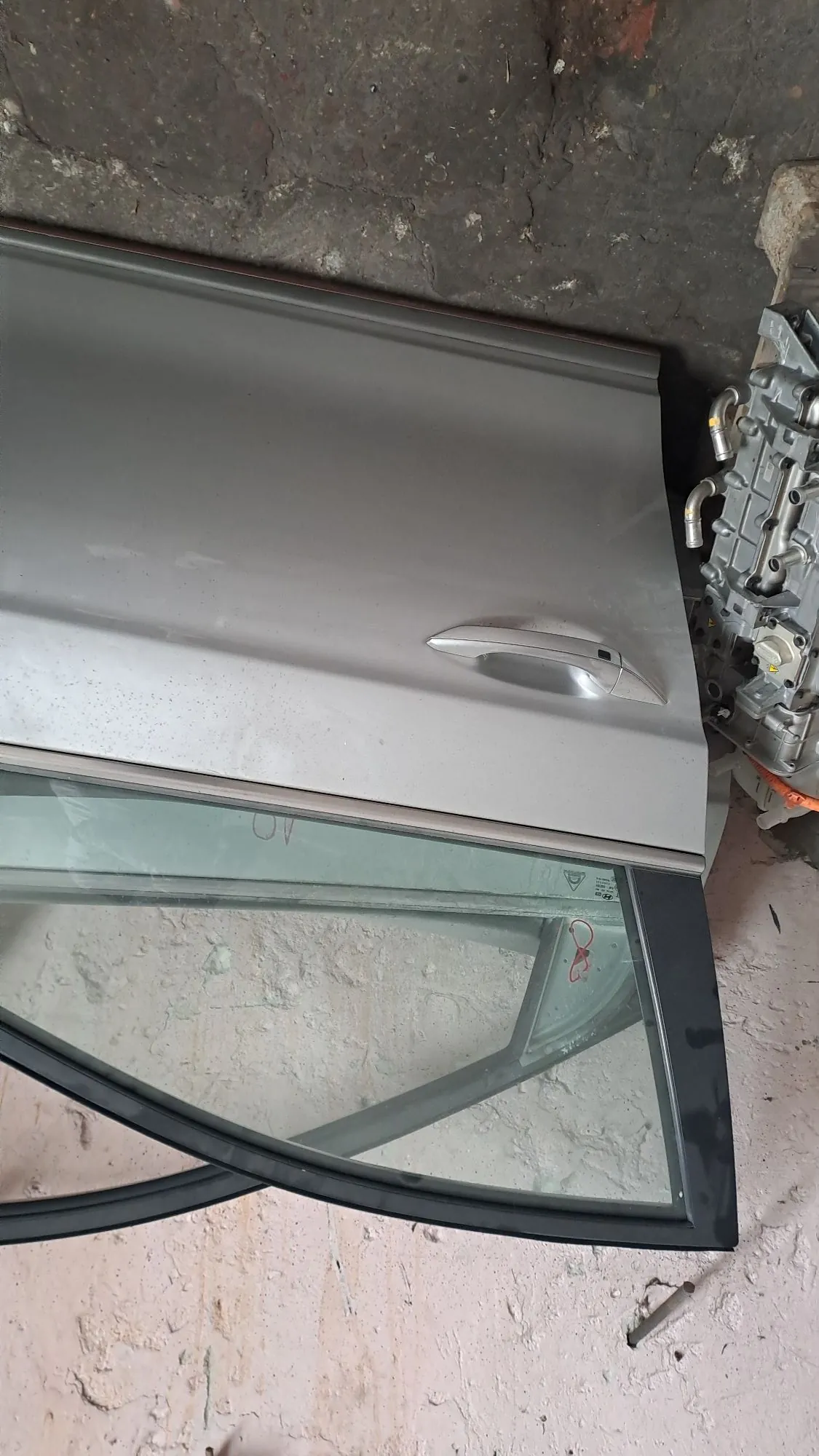 Srebrne drzwi samochodowe z szybą, oparte o jasną, chropowatą ścianę. Drzwi są ułożone poziomo, a pod nimi widoczny jest fragment silnika lub podzespołów mechanicznych.