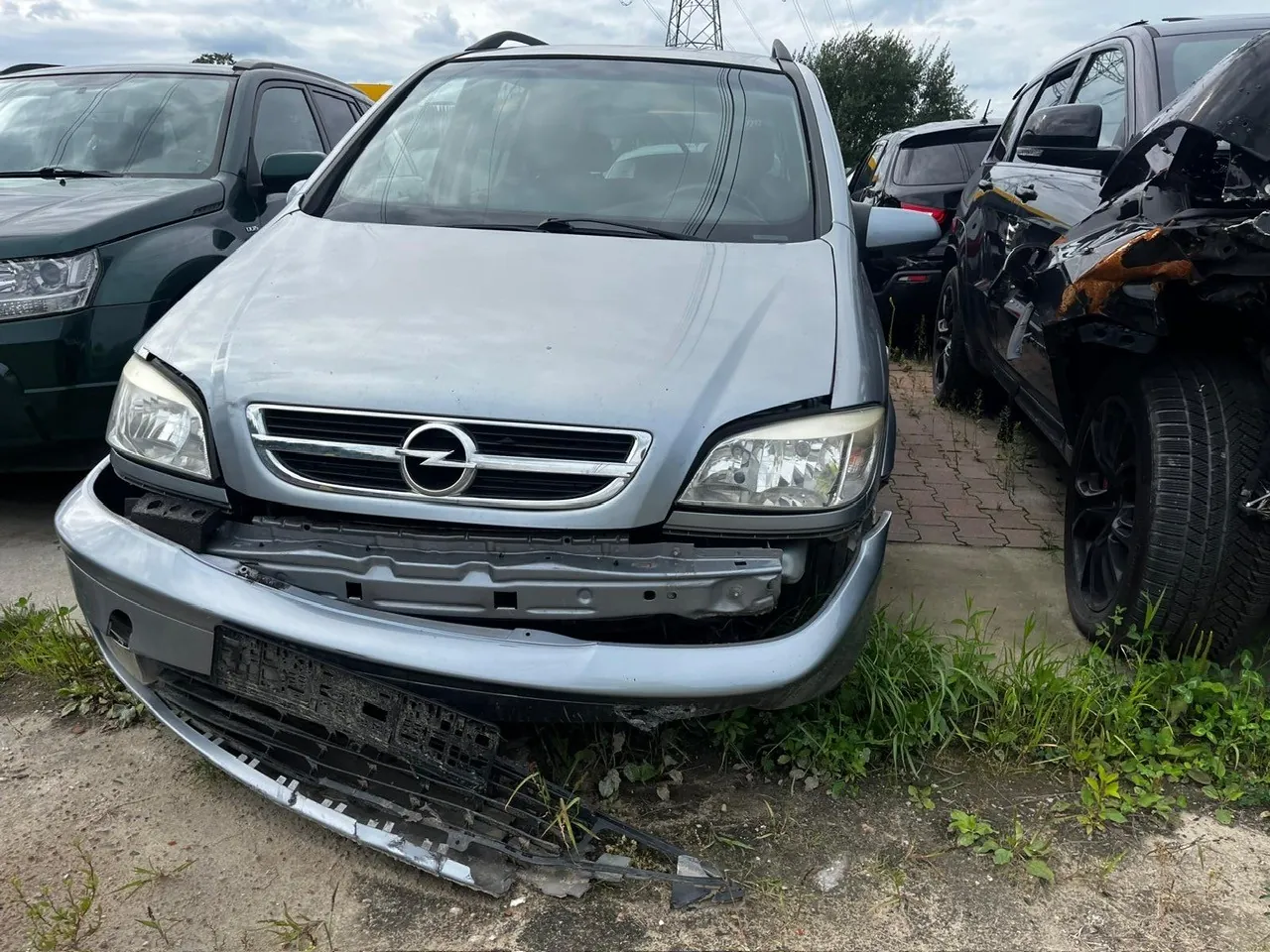 Srebrny samochód marki Opel z uszkodzonym przednim zderzakiem, który częściowo odpadł i leży na ziemi. Pojazd znajduje się na parkingu obok innych samochodów, w tym ciemnozielonego SUV-a po lewej i czarnego, mocno rozbitego samochodu po prawej.