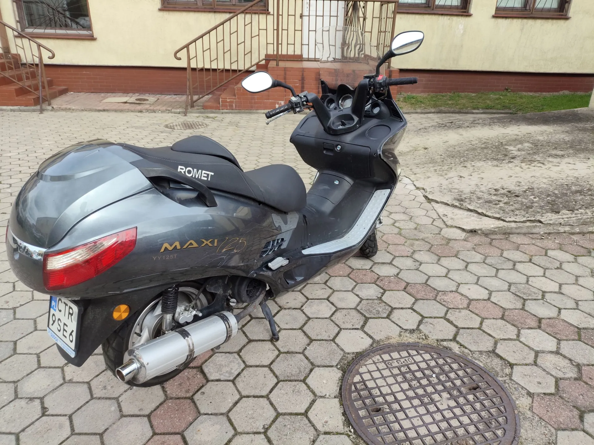 Szary skuter marki Romet Maxi 125 zaparkowany na kostce brukowej. Pojazd ma tablicę rejestracyjną o numerze CTR 9SE6. W tle widoczny jest fragment budynku z schodami i metalową balustradą oraz okna. Na ziemi obok skutera znajduje się okrągły właz kanalizacyjny.