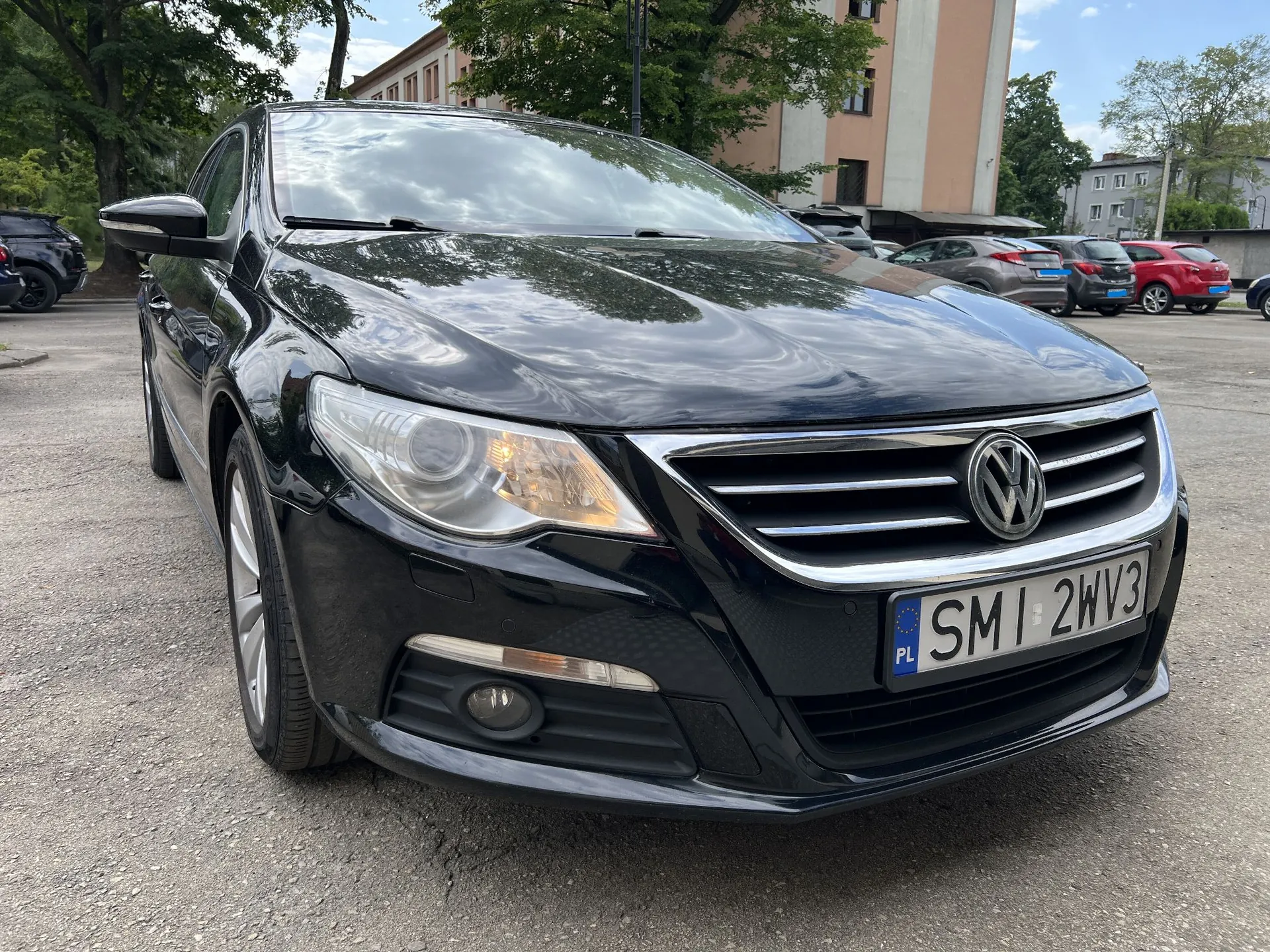 Czarny samochód osobowy marki Volkswagen zaparkowany na asfaltowym placu. Widoczny jest przód pojazdu z rejestracją SMI 2WV3. W tle znajdują się inne zaparkowane samochody, drzewa oraz wielopiętrowy budynek.