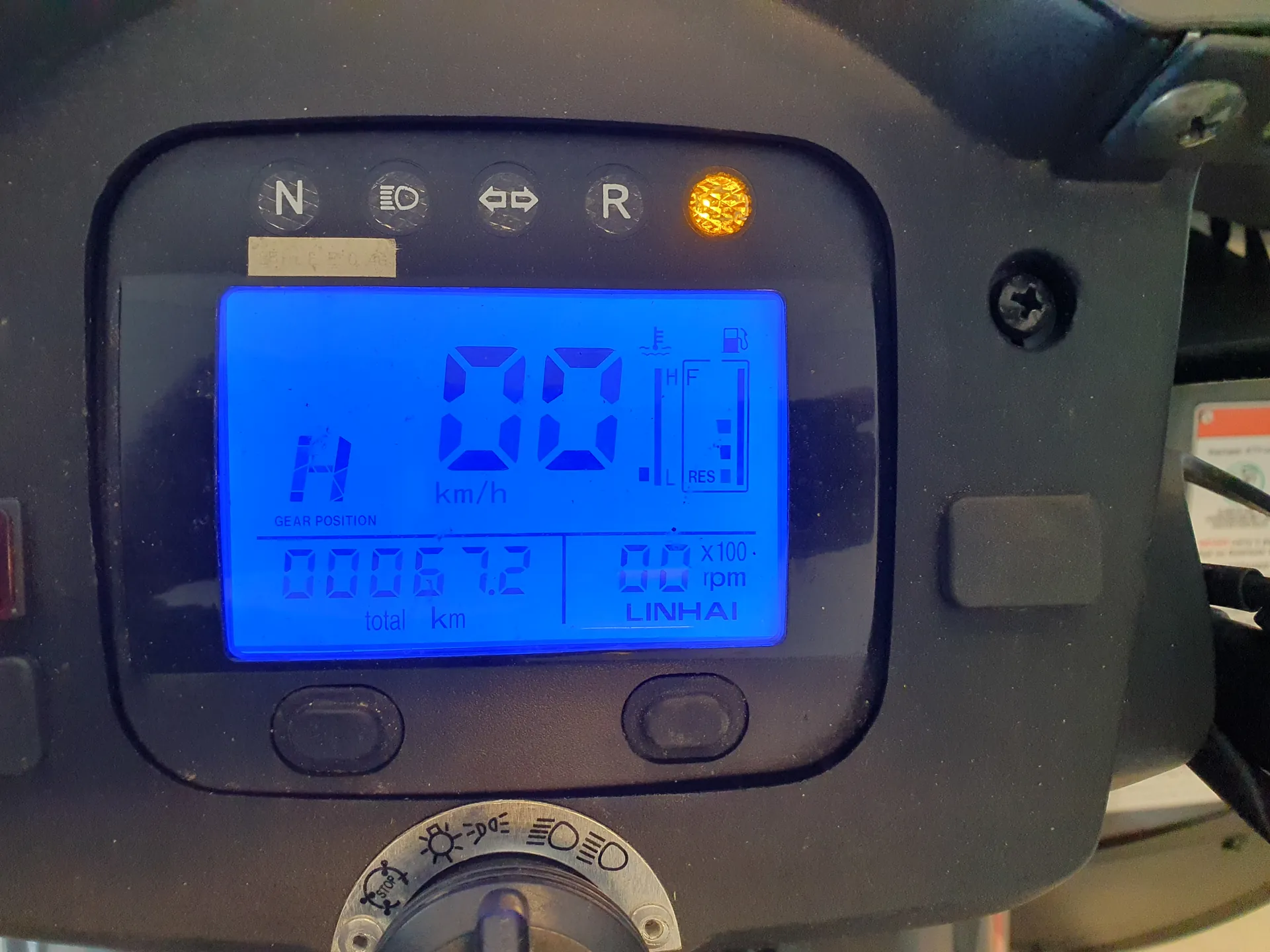 Bliski widok czarnego panelu deski rozdzielczej pojazdu z niebieskim wyświetlaczem cyfrowym. Wyświetlacz pokazuje prędkość 00 km/h, pozycję biegu H (podpisane 'GEAR POSITION'), przebieg 00067.2 km (podpisane 'total km'), obroty 00 x100 rpm oraz nazwę producenta 'LINHAI'. W górnej części panelu widoczne są wskaźniki: N, symbol reflektora, kierunkowskazy, R oraz świecąca pomarańczowa dioda ostrzegawcza. Na wyświetlaczu znajduje się również wskaźnik paliwa z oznaczeniami H, L i RES. Poniżej wyświetlacza znajdują się dwa przyciski i pokrętło z symbolami oświetlenia.