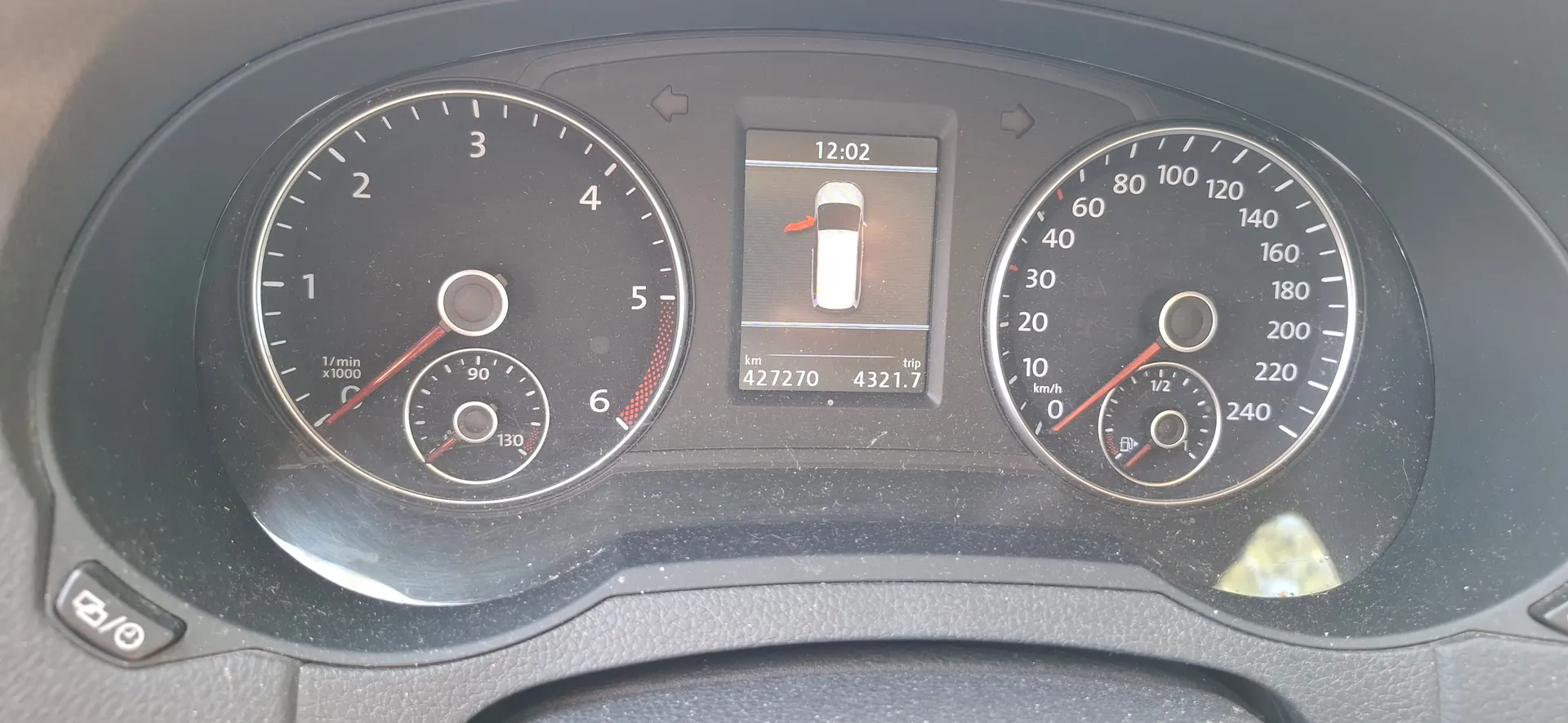 Panel instrumentów samochodu. Po lewej stronie znajduje się obrotomierz z zaznaczonymi wartościami od 1 do 6 (x1000 1/min) oraz mniejszy wskaźnik temperatury cieczy chłodzącej. W centrum znajduje się cyfrowy wyświetlacz pokazujący godzinę 12:02, ikonę poziomu oleju oraz przebieg całkowity 427270 km i przebieg podróży 4321.7 km. Po prawej stronie znajduje się prędkościomierz z podziałką od 0 do 240 km/h oraz wskaźnik poziomu paliwa.