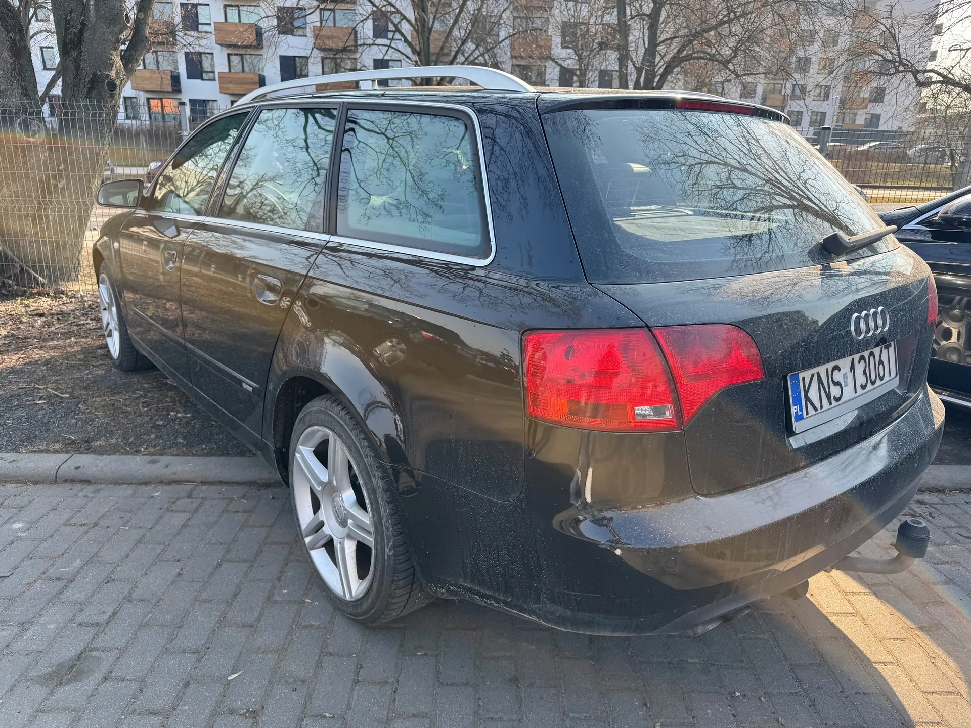 Czarny samochód Audi typu kombi zaparkowany na brukowanej powierzchni. Widok od tyłu z widocznym logo Audi, czerwonymi światłami tylnymi oraz tablicą rejestracyjną KNS 1306T z niebieskim pasem PL po lewej stronie. Samochód posiada srebrny dachówkarz i felgi. W tle budynek mieszkalny z białymi i brązowymi elewacjami, drzewa bez liści, metalowe ogrodzenie oraz fragment innego czarnego pojazdu po prawej stronie. Podłoga składa się z bruku i częściowo z ziemi z suchą trawą.