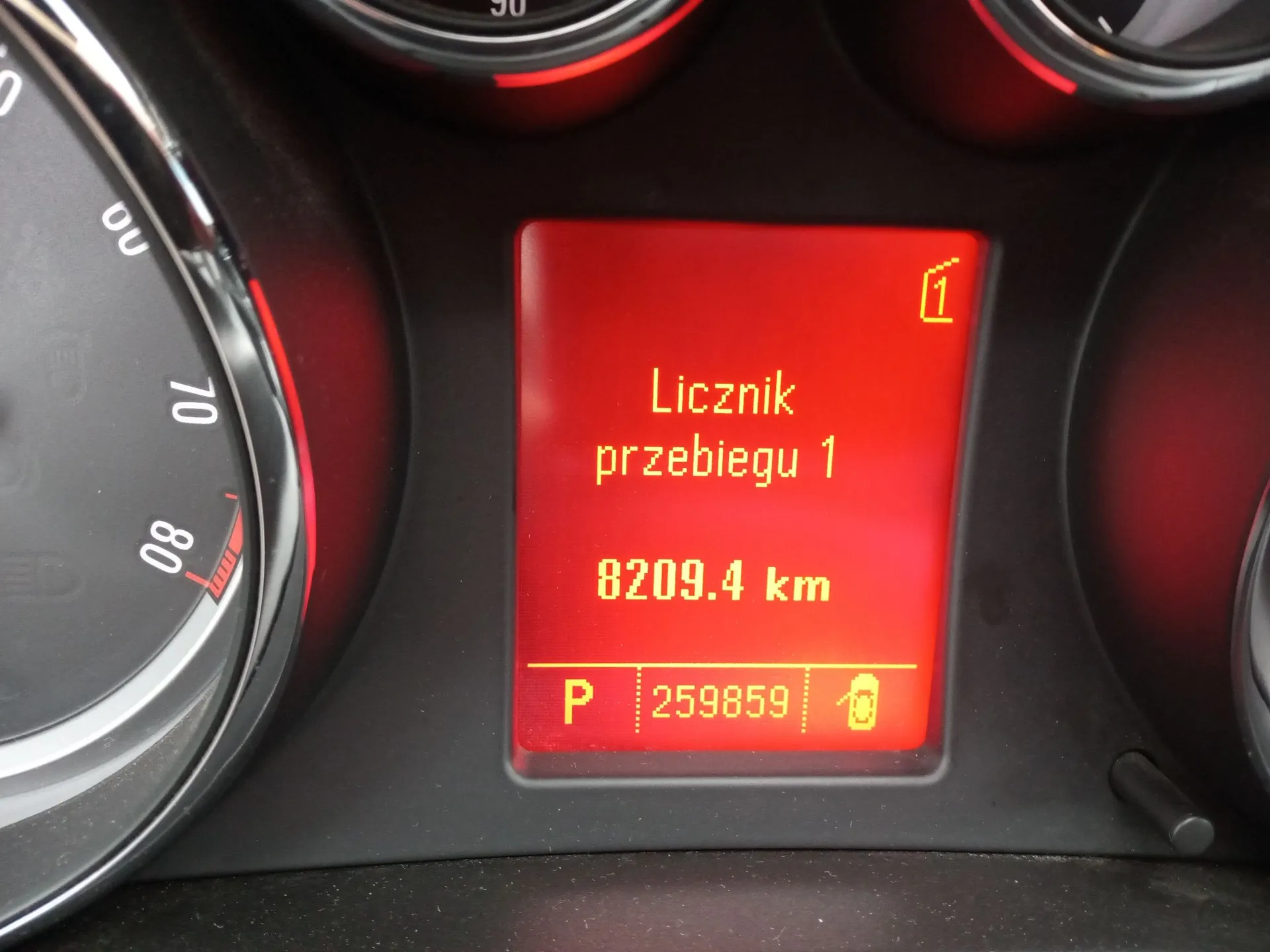 Zbliżenie na czerwony wyświetlacz cyfrowy w desce rozdzielczej samochodu. Na ekranie widnieje napis „Licznik przebiegu 1”, poniżej wartość „8209.4 km”, a na dole w oddzielnej sekcji litera „P”, liczba „259859” oraz ikona zamkniętej kłódki. Po lewej stronie widoczny jest fragment analogowego prędkościomierza z oznaczeniami 70 i 80.